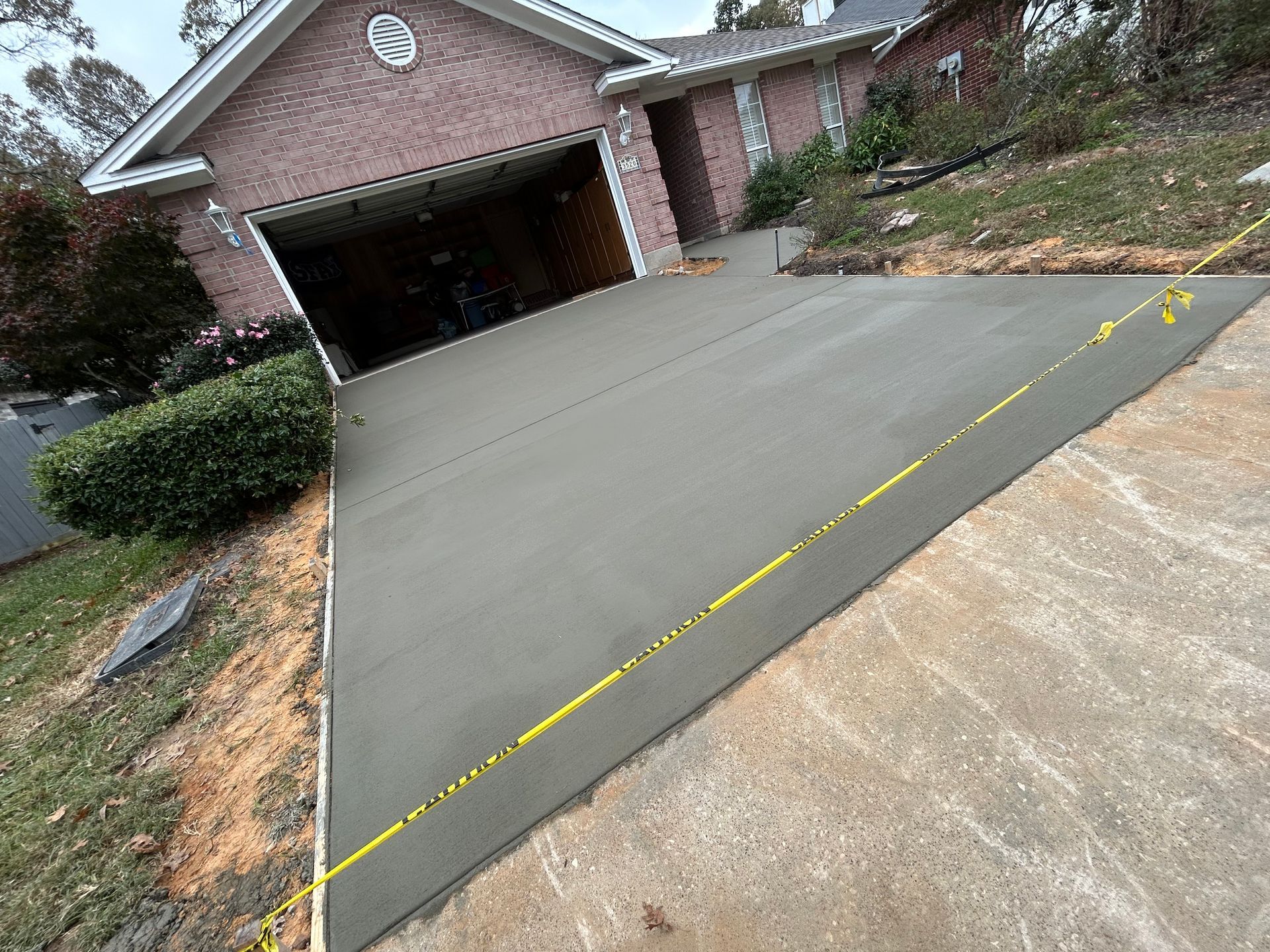 A newly poured, smooth gray concrete driveway in front of a brick house, cordoned off with yellow caution tape.