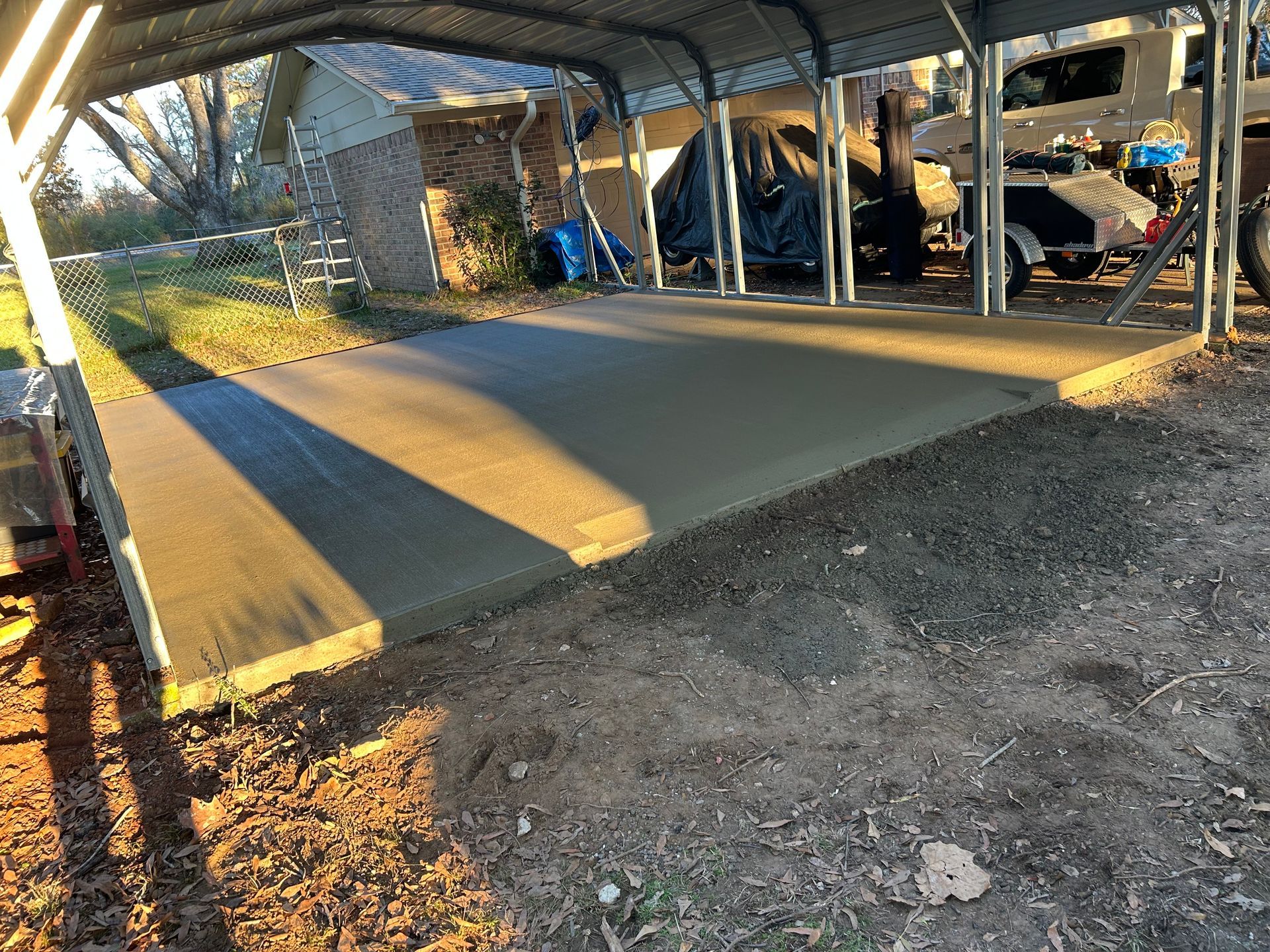 A newly poured concrete slab sits under a metal carport structure, situated next to a house with a dirt-covered yard.