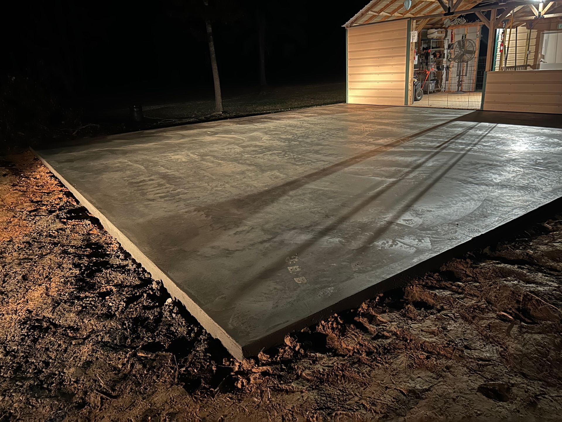 Freshly poured, wet concrete patio slab at night, situated in front of a partially constructed wooden building.