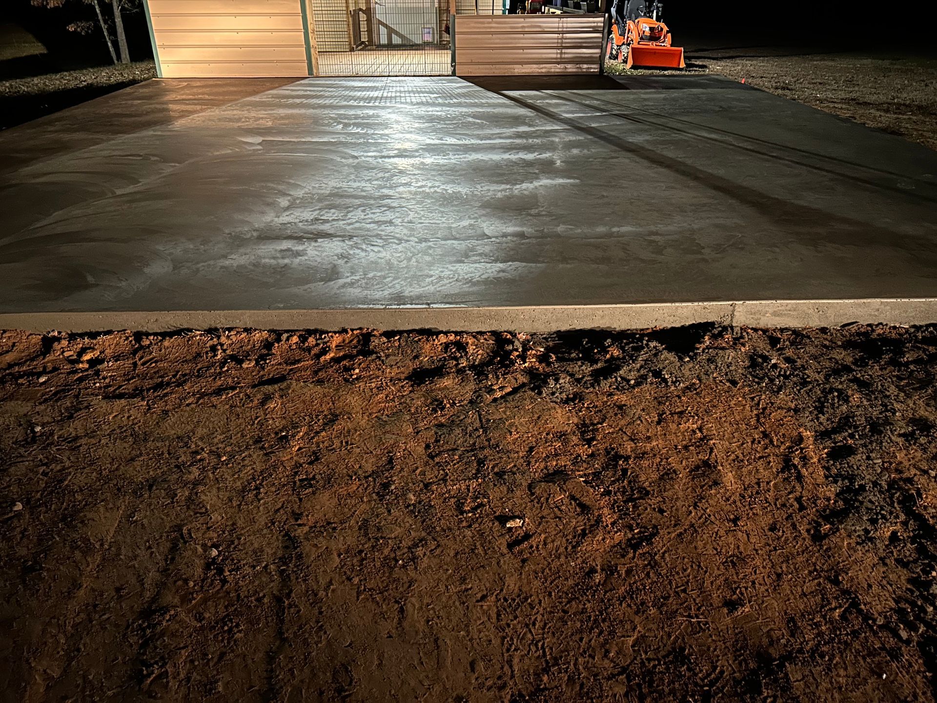Freshly poured concrete slab in front of a building at night, with dark soil in the foreground.