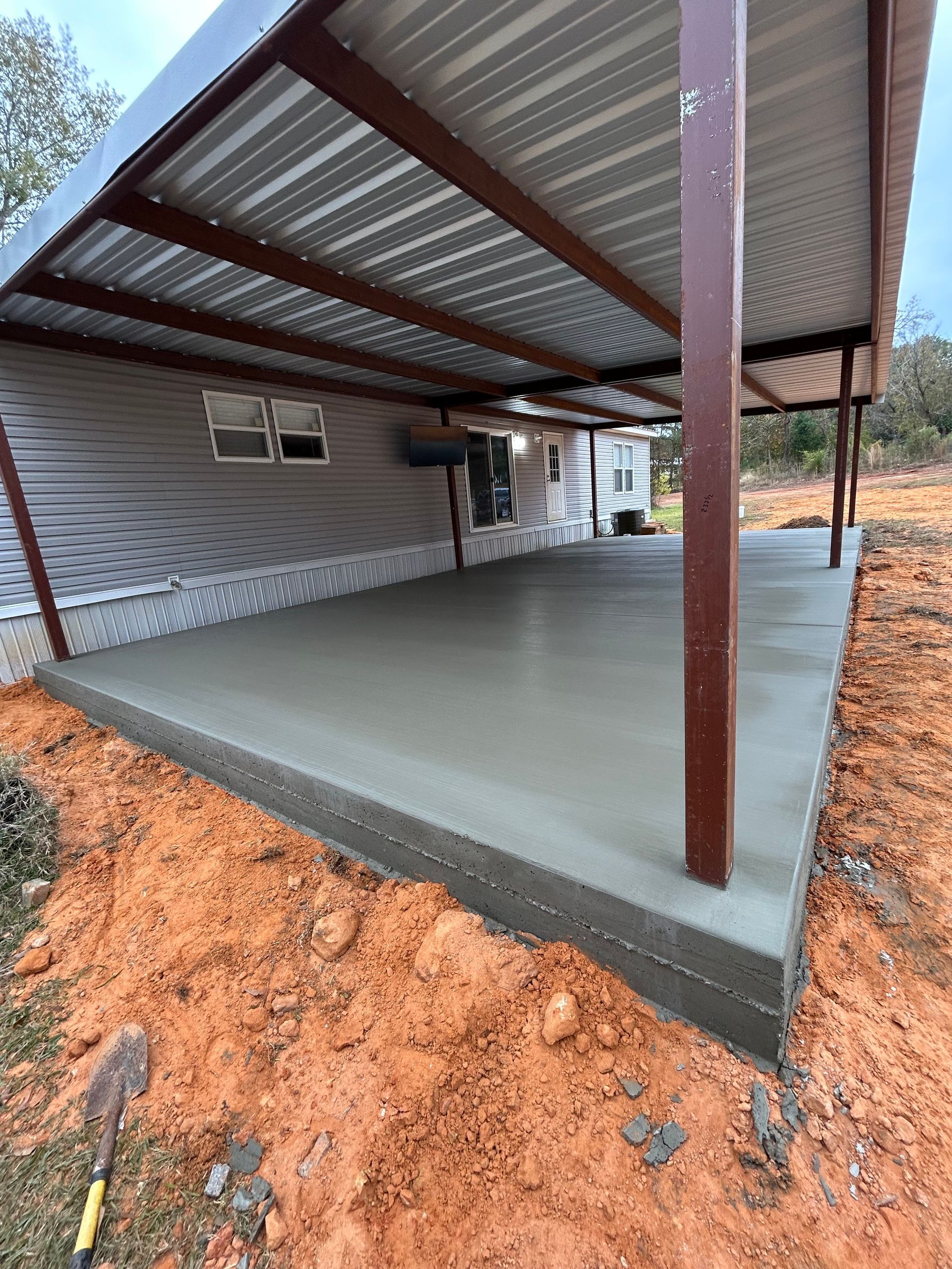 Freshly poured concrete patio slab under a metal carport roof attached to the side of a house with red dirt surroundings.