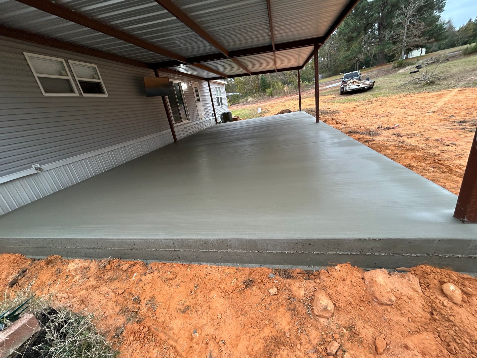 A fresh, smooth concrete patio slab poured under a metal carport attached to the side of a light gray house.
