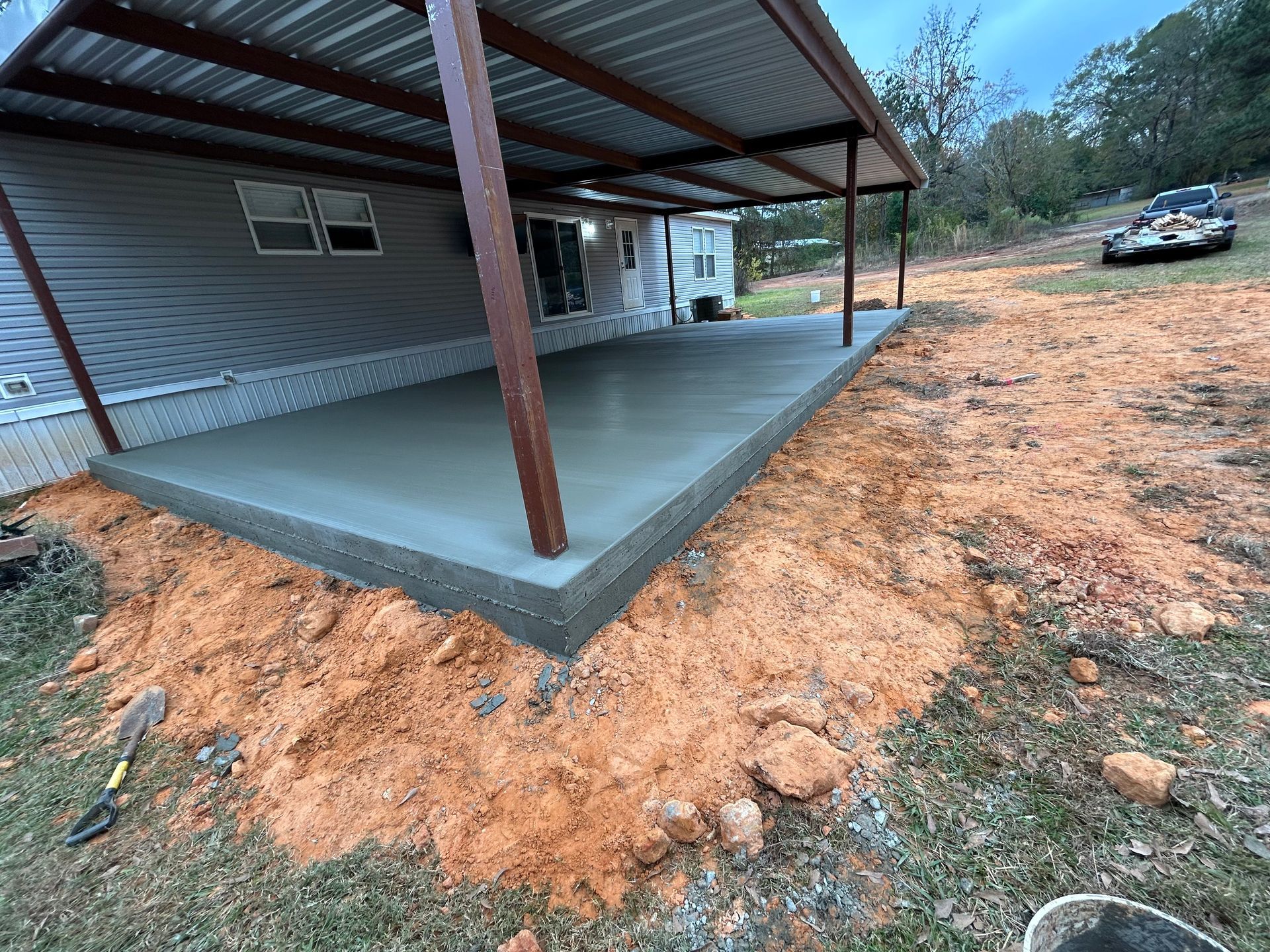 A newly poured concrete patio slab underneath a metal roof structure attached to a gray siding home with dirt surroundings.