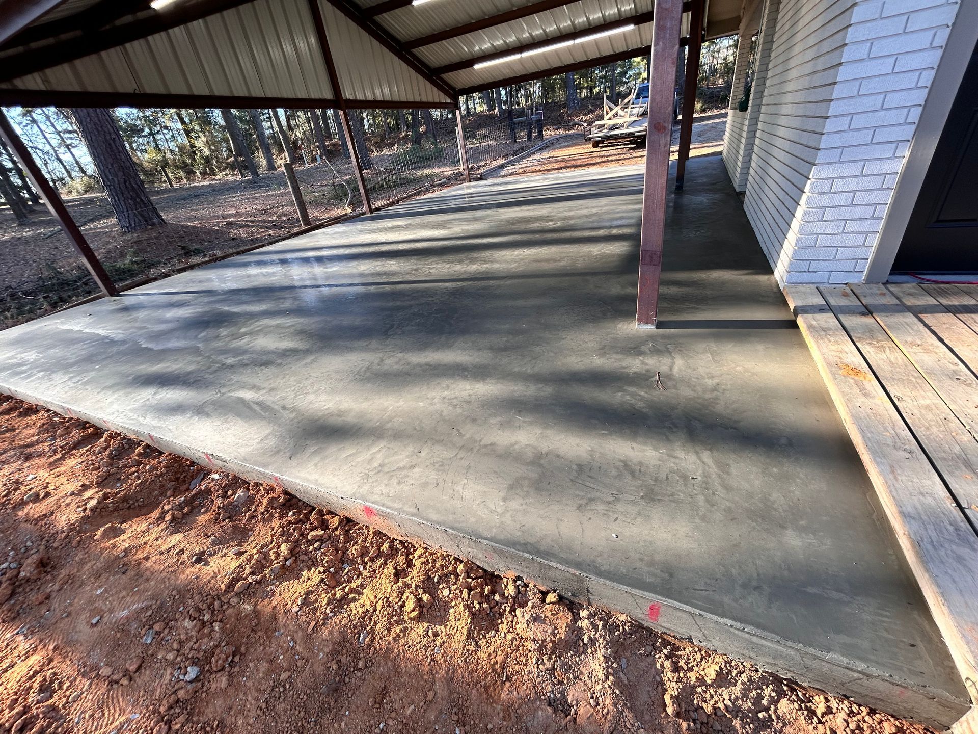 A freshly poured concrete patio under a covered carport attached to a white brick house, bordered by bare dirt.