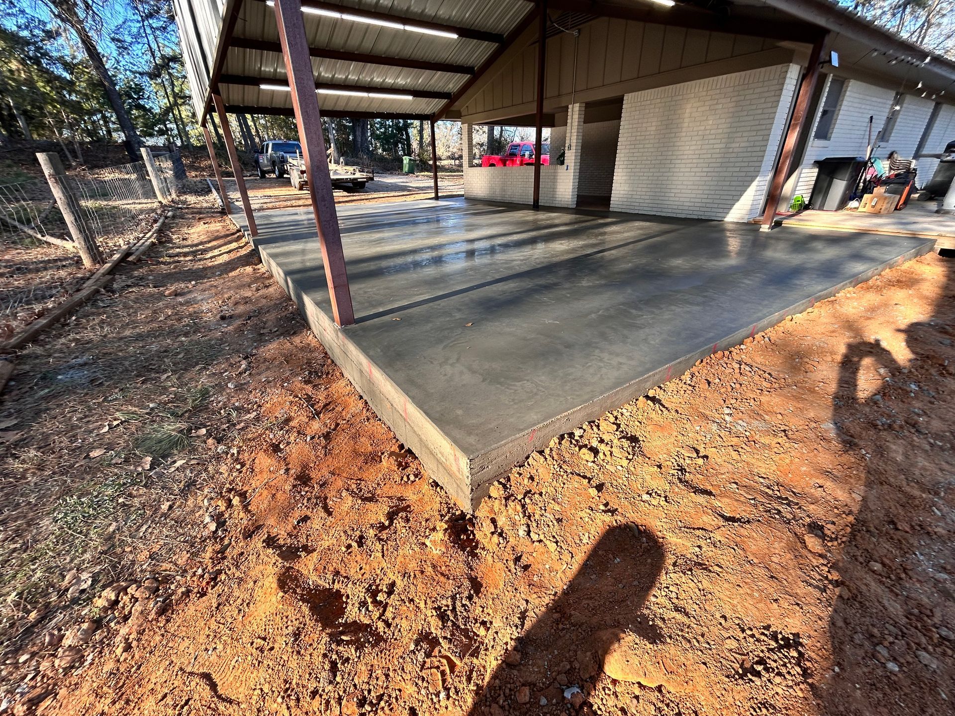 A newly poured concrete driveway slab sits under a metal carport roof beside a house, surrounded by red dirt.