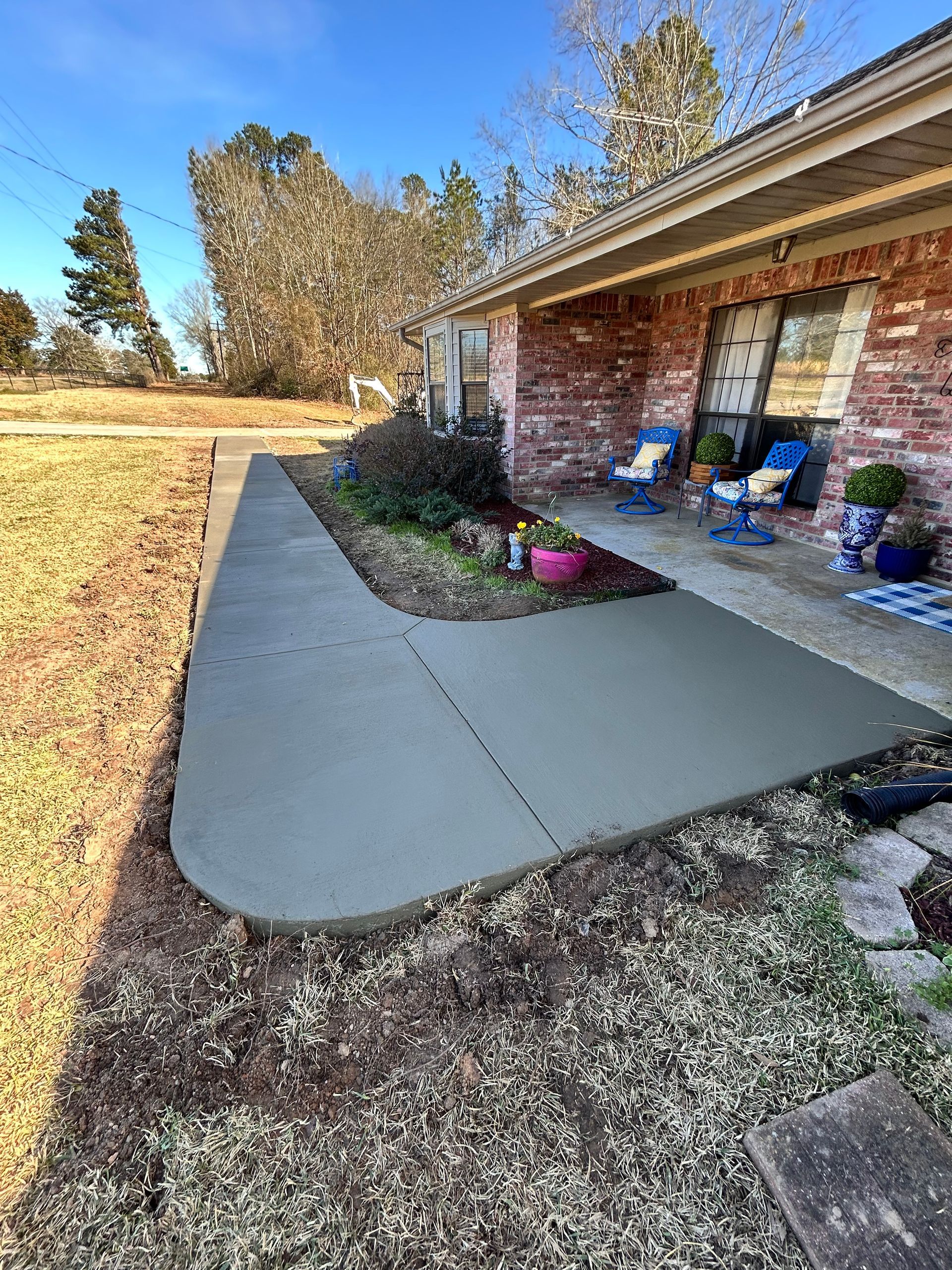 A newly poured concrete walkway curves in front of a brick house with a small patio and potted plants.