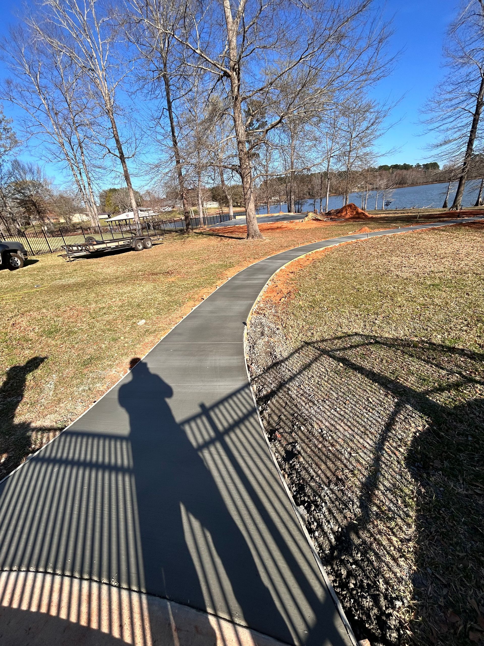 A curved concrete walkway leads through a sunny outdoor area toward a body of water, with a person’s shadow in the foreground.
