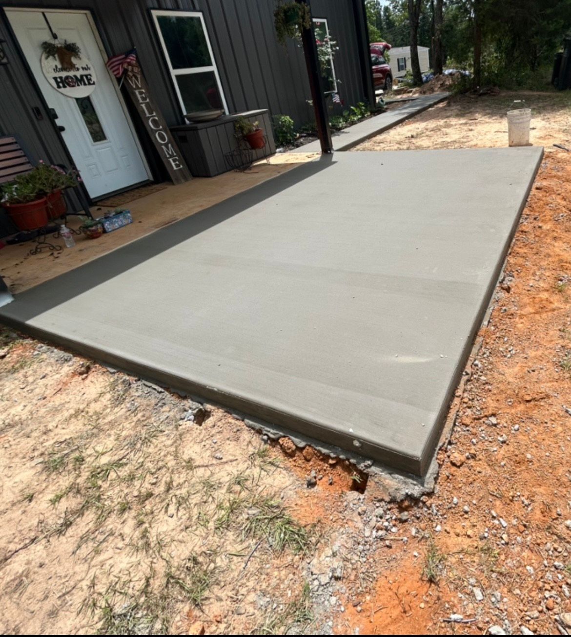 A newly poured concrete patio slab outside a dark gray house with a white front door and a welcome sign.