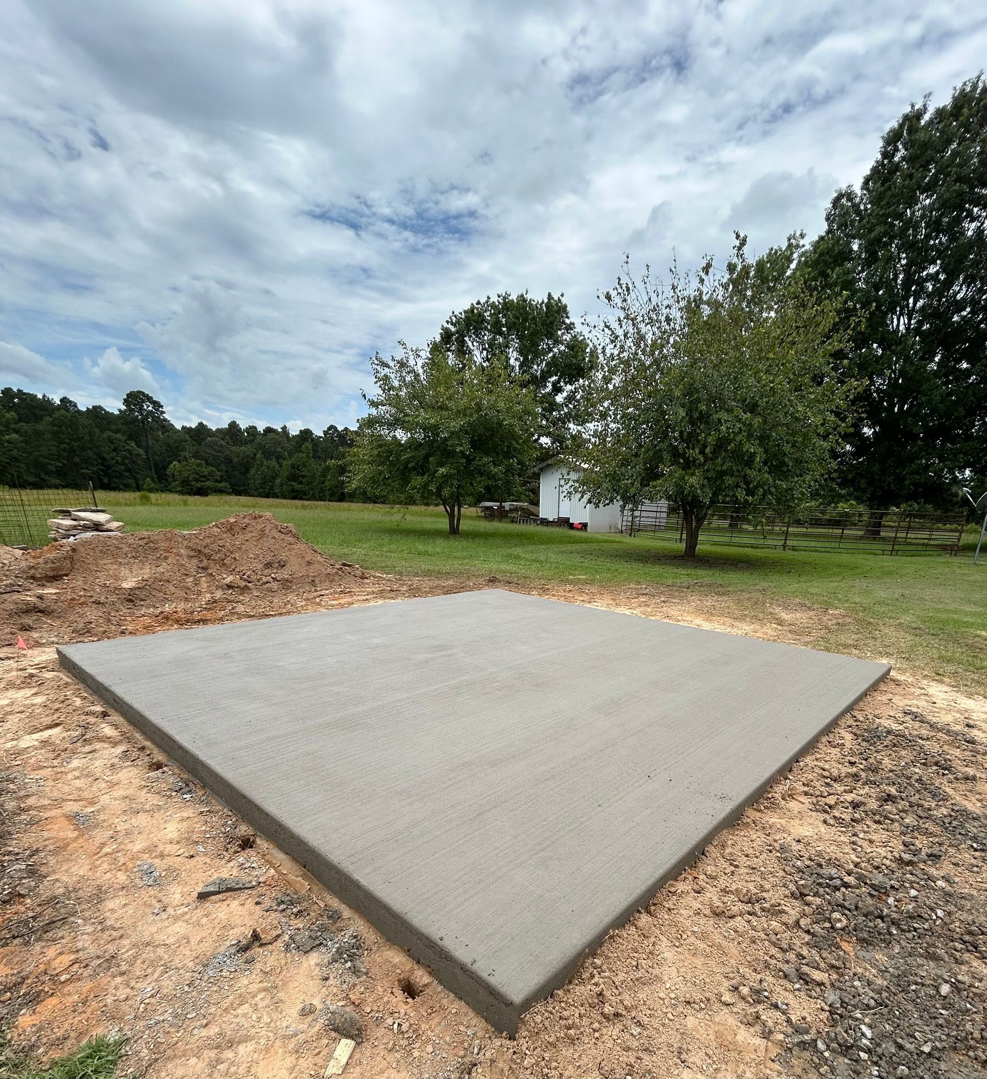 A freshly poured concrete slab set in a grassy field with trees and a white shed in the distance.
