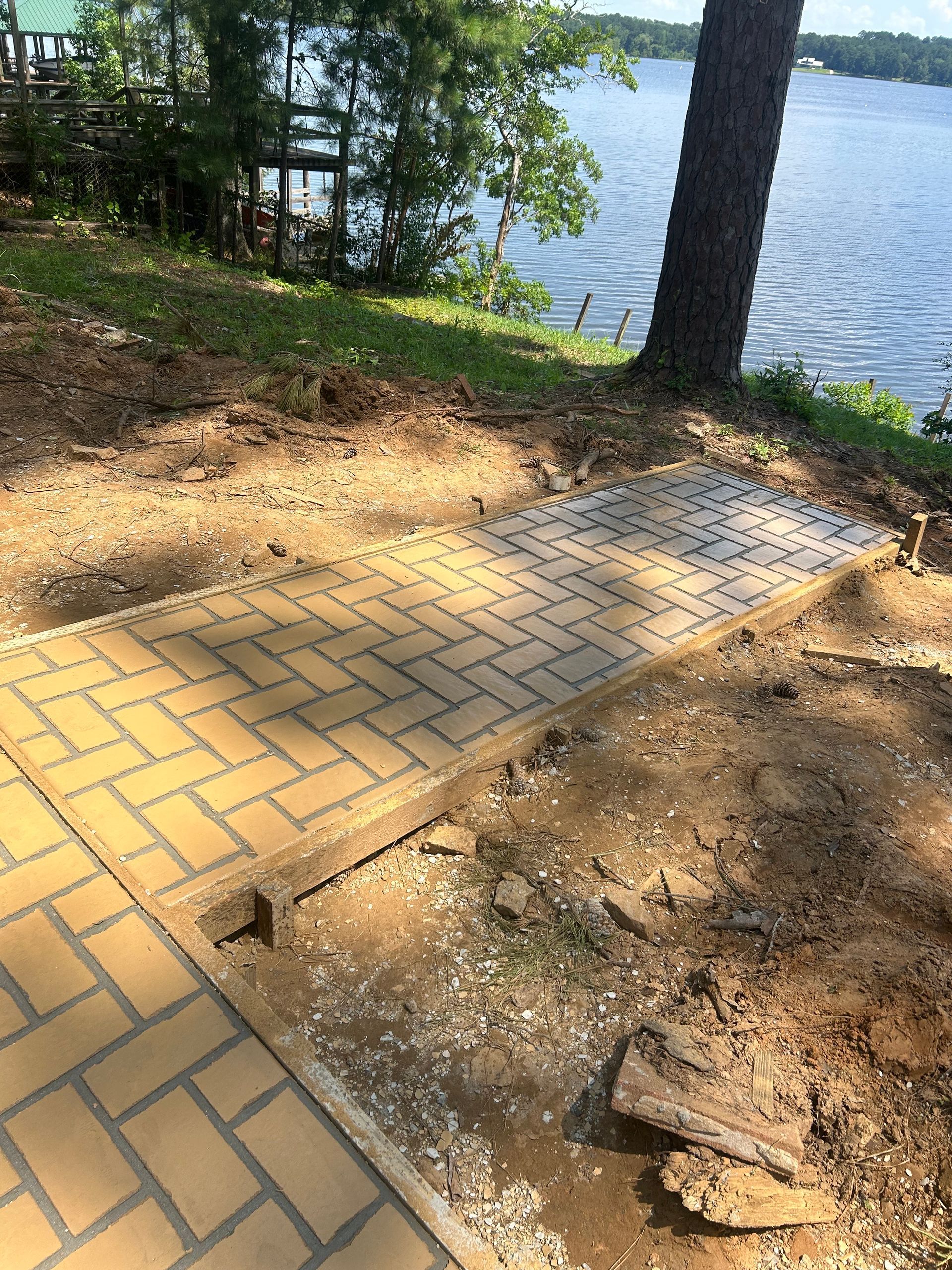 A tan brick walkway in a herringbone pattern leads toward a lake surrounded by trees and a wooden deck.