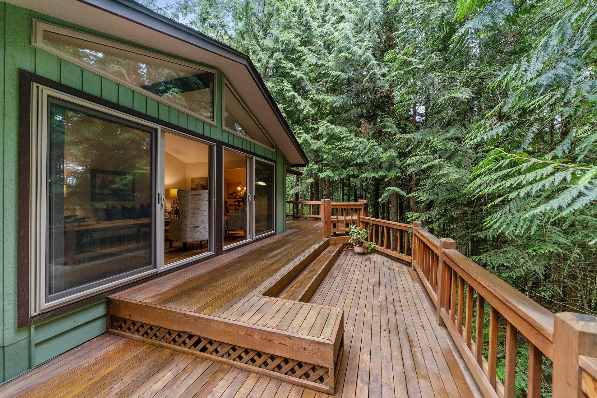 Wooden deck of a green house surrounded by trees, with large windows.