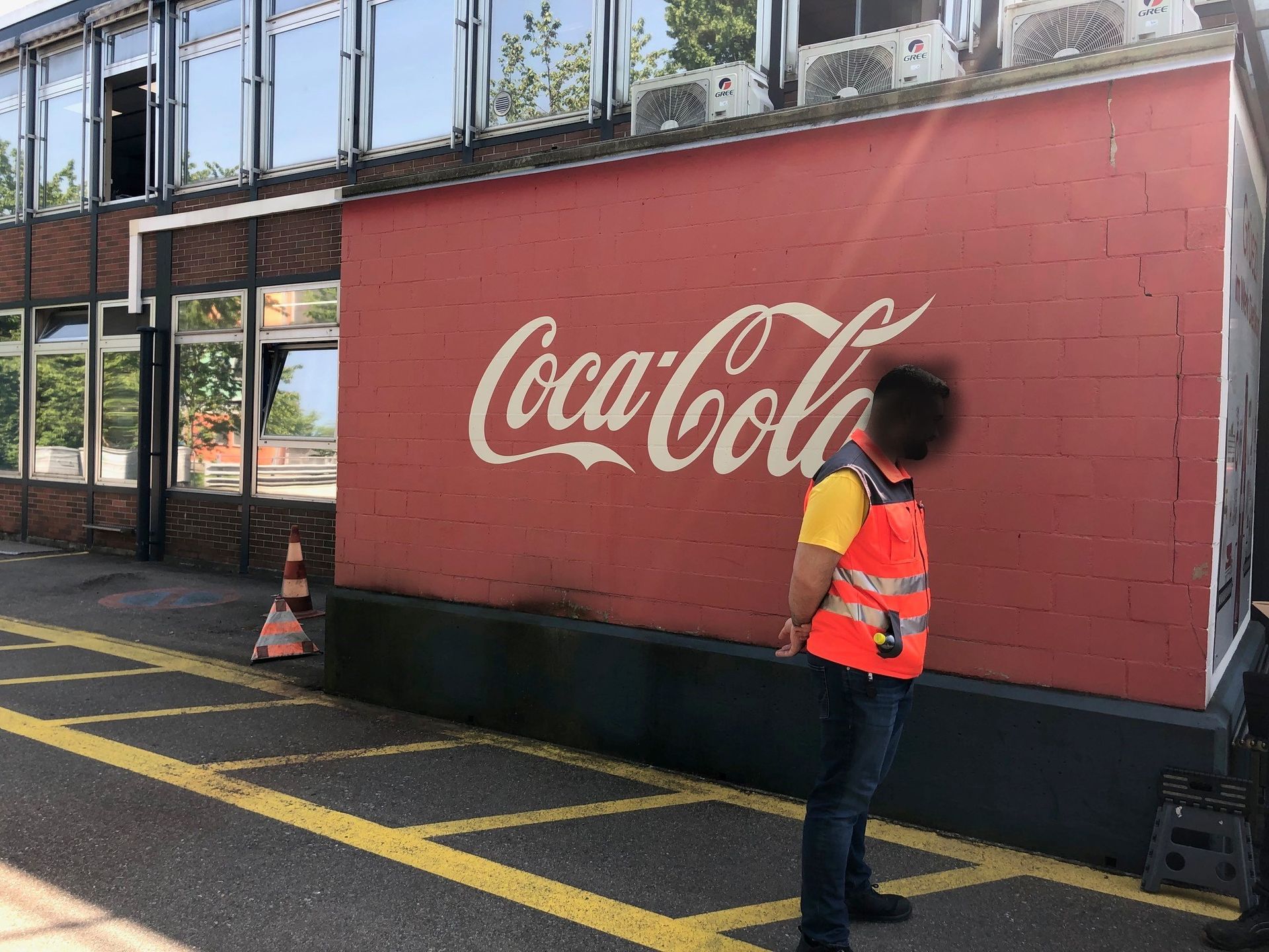 Ein Mann in einer orangefarbenen Weste steht vor einem Coca-Cola-Schild an einer roten Wand.