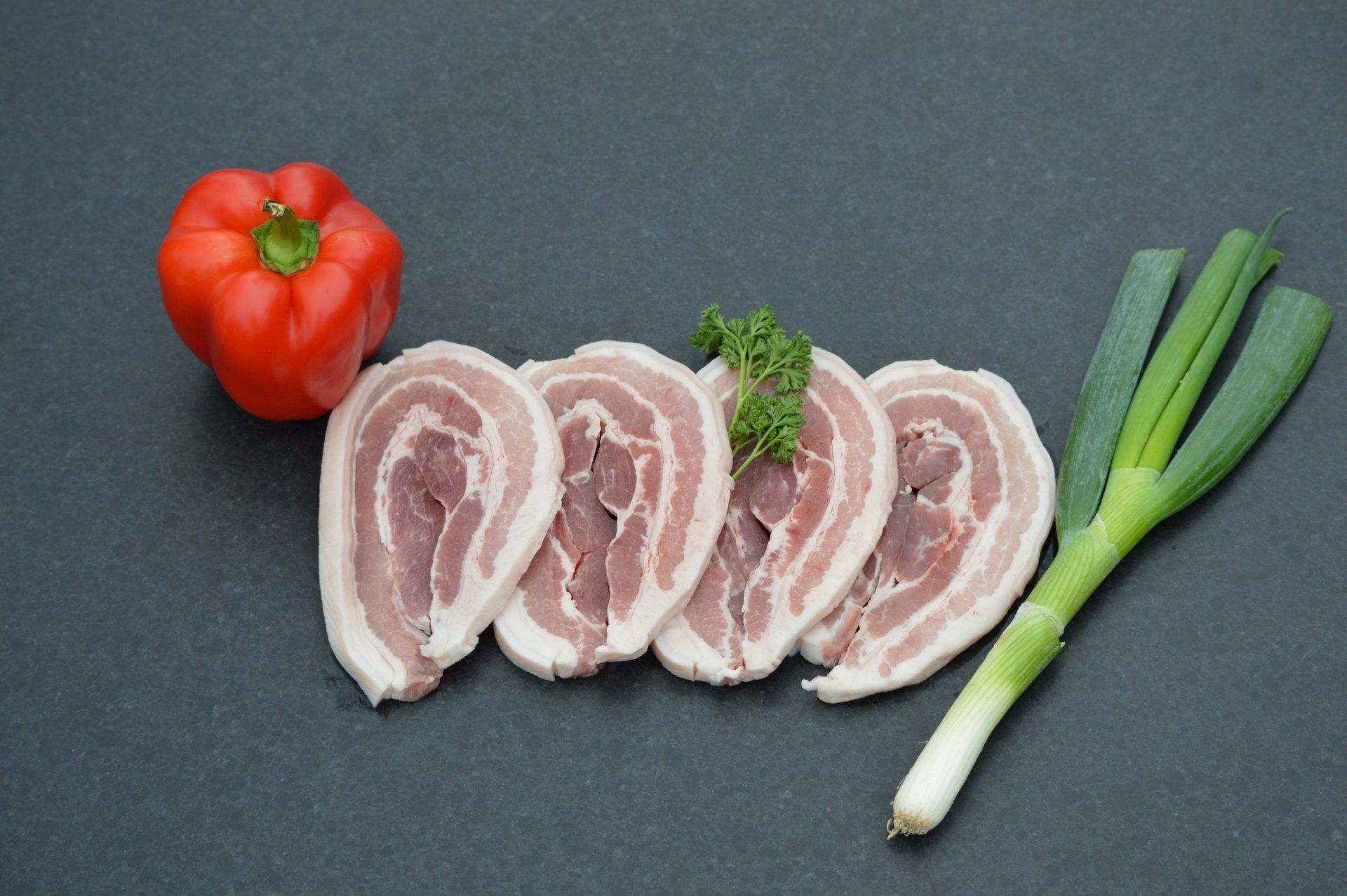 Gesneden spek met rode paprika, groene uien en peterselie op een donker oppervlak.