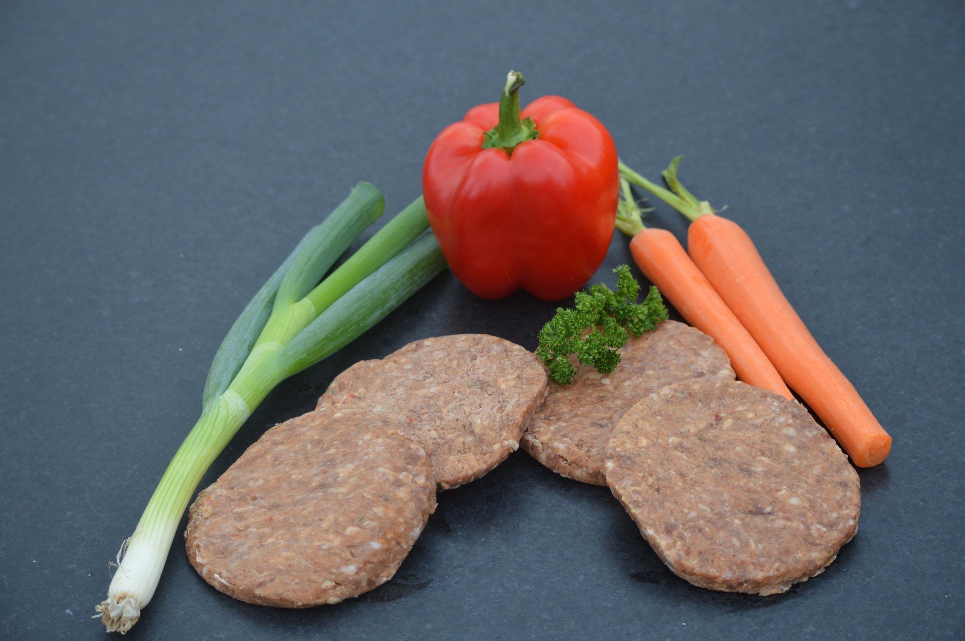 Ronde, bruine burgers met rode paprika, wortels, groene uien en peterselie op een donkere ondergrond.