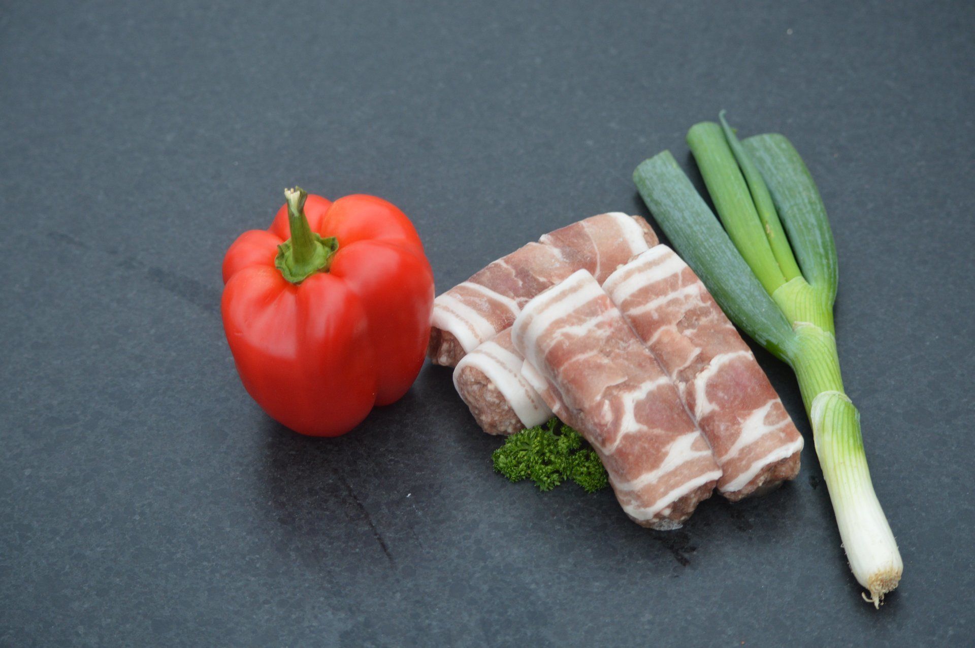 Rode paprika, in spek gewikkelde vleesrolletjes en groene uien op een donker oppervlak.