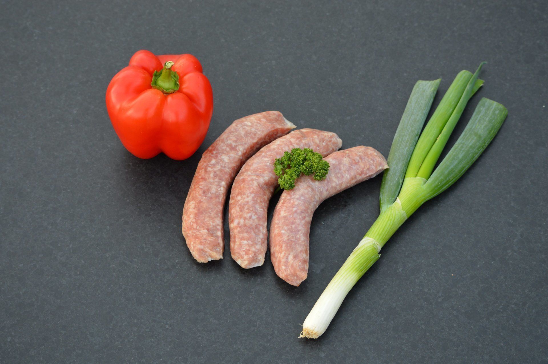 Drie worstjes, rode paprika en groene ui op een donker oppervlak.