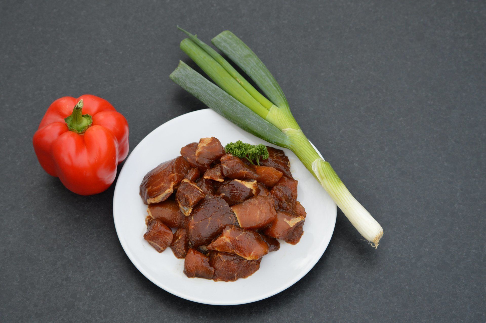Gebakken vlees op een bord met rode paprika en groene uien op een donker oppervlak.