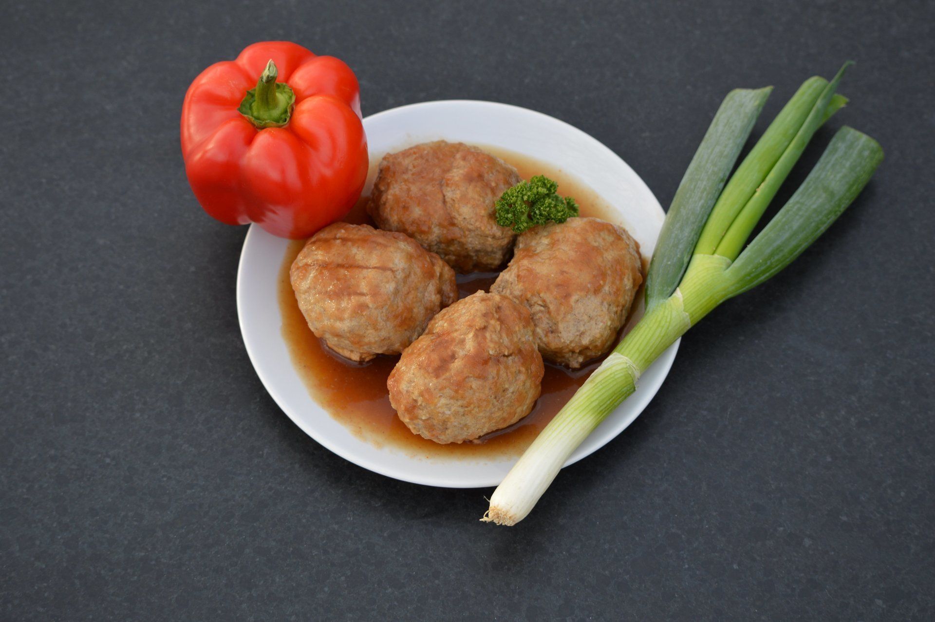 Een bord met vier ronde, bruine gehaktballetjes in bouillon, met rode paprika en groene ui.