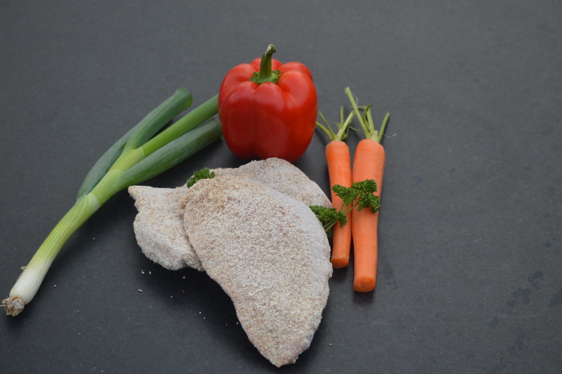 Gepaneerde kipfilets, rode paprika, wortels en bosuitjes op een zwart oppervlak.