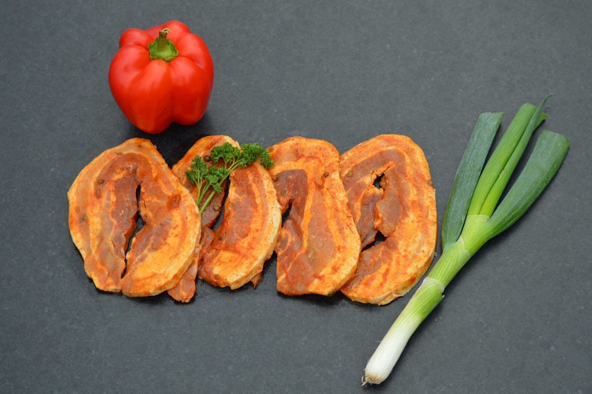 Gesneden, gekruid vlees met rode paprika en groene ui op een donker oppervlak.