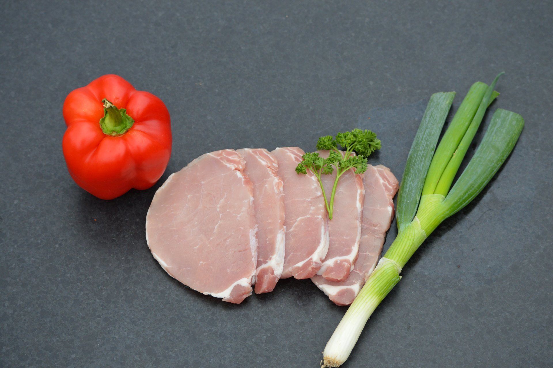 Rauwe varkenskoteletten, rode paprika en groene uien op een donker oppervlak.