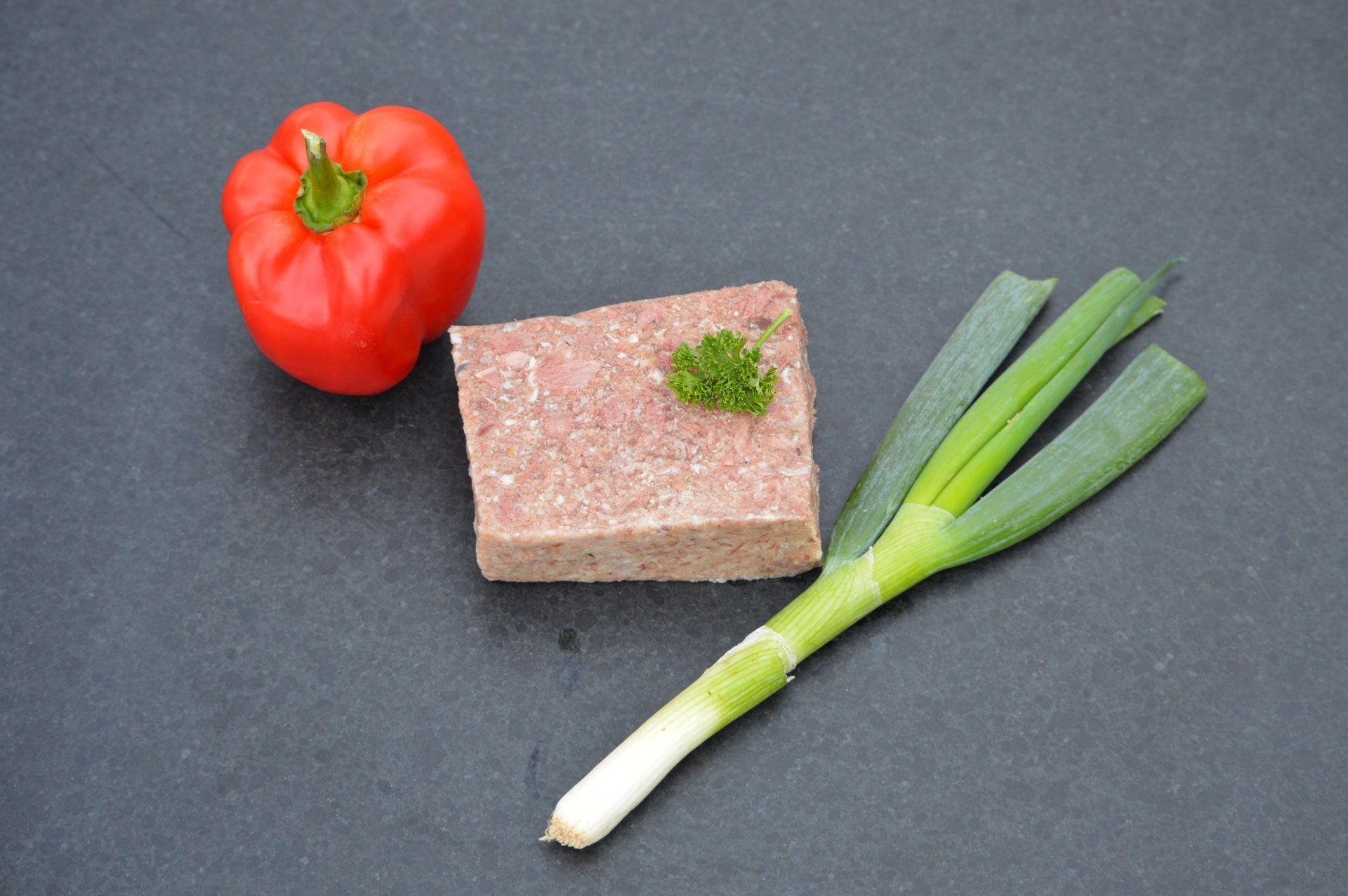 Rode paprika, een blokje vlees met peterselie en een lente-ui op een donkere ondergrond.