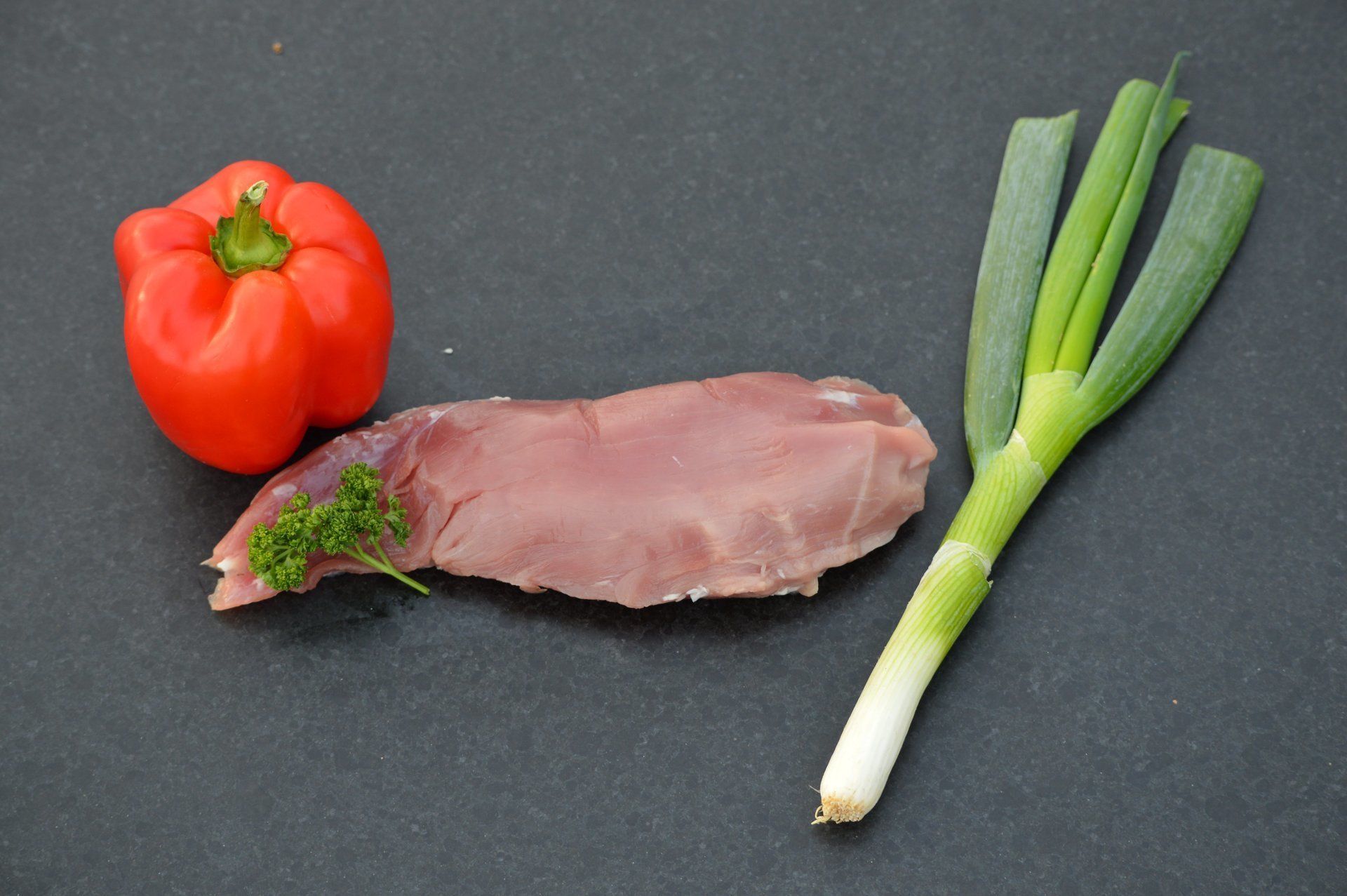 Rode paprika, rauw vlees, groene ui en peterselie op een donker oppervlak.