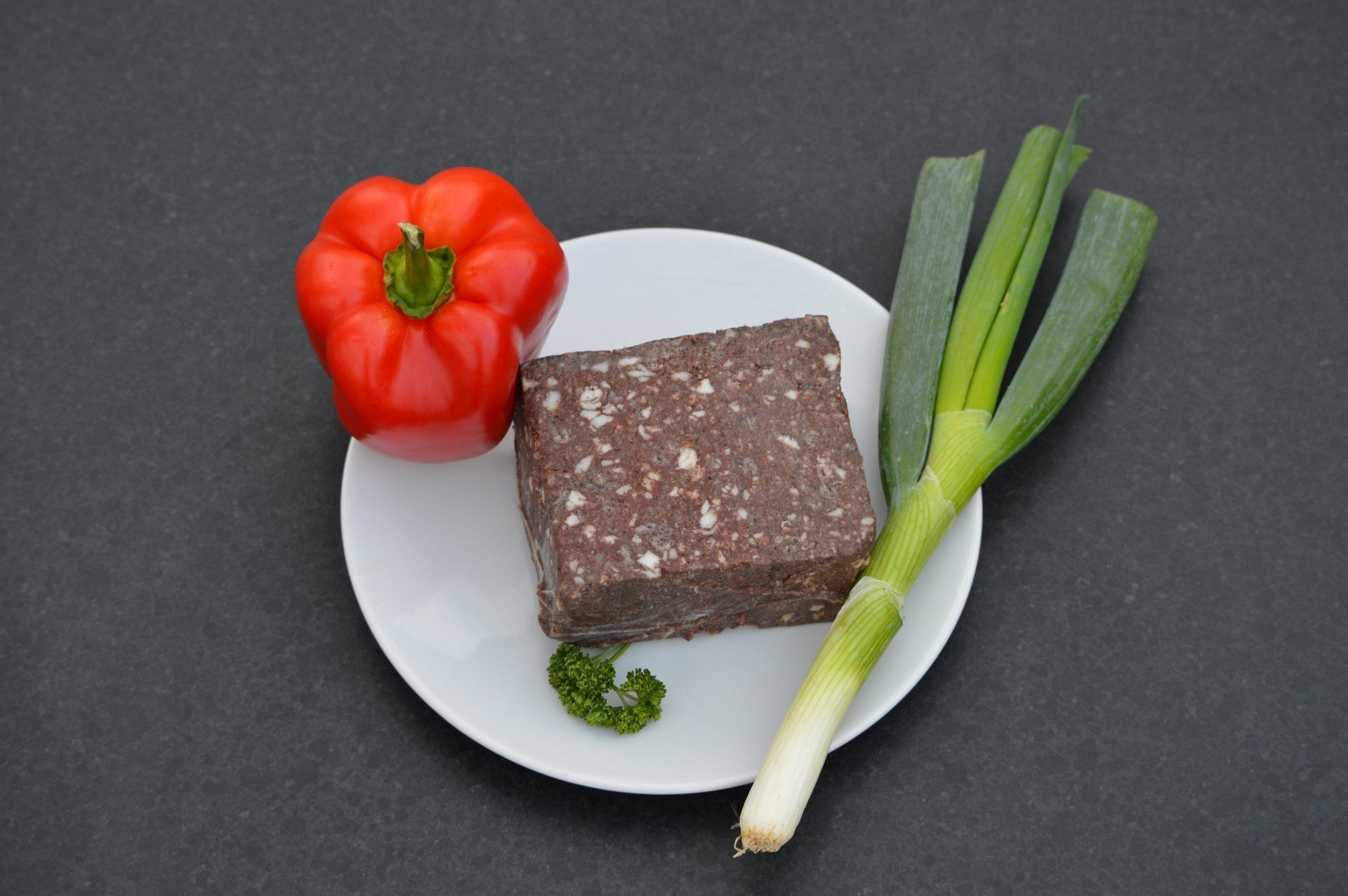 Blok bloedworst op een bord met rode paprika, prei en peterselie op een donkere achtergrond.
