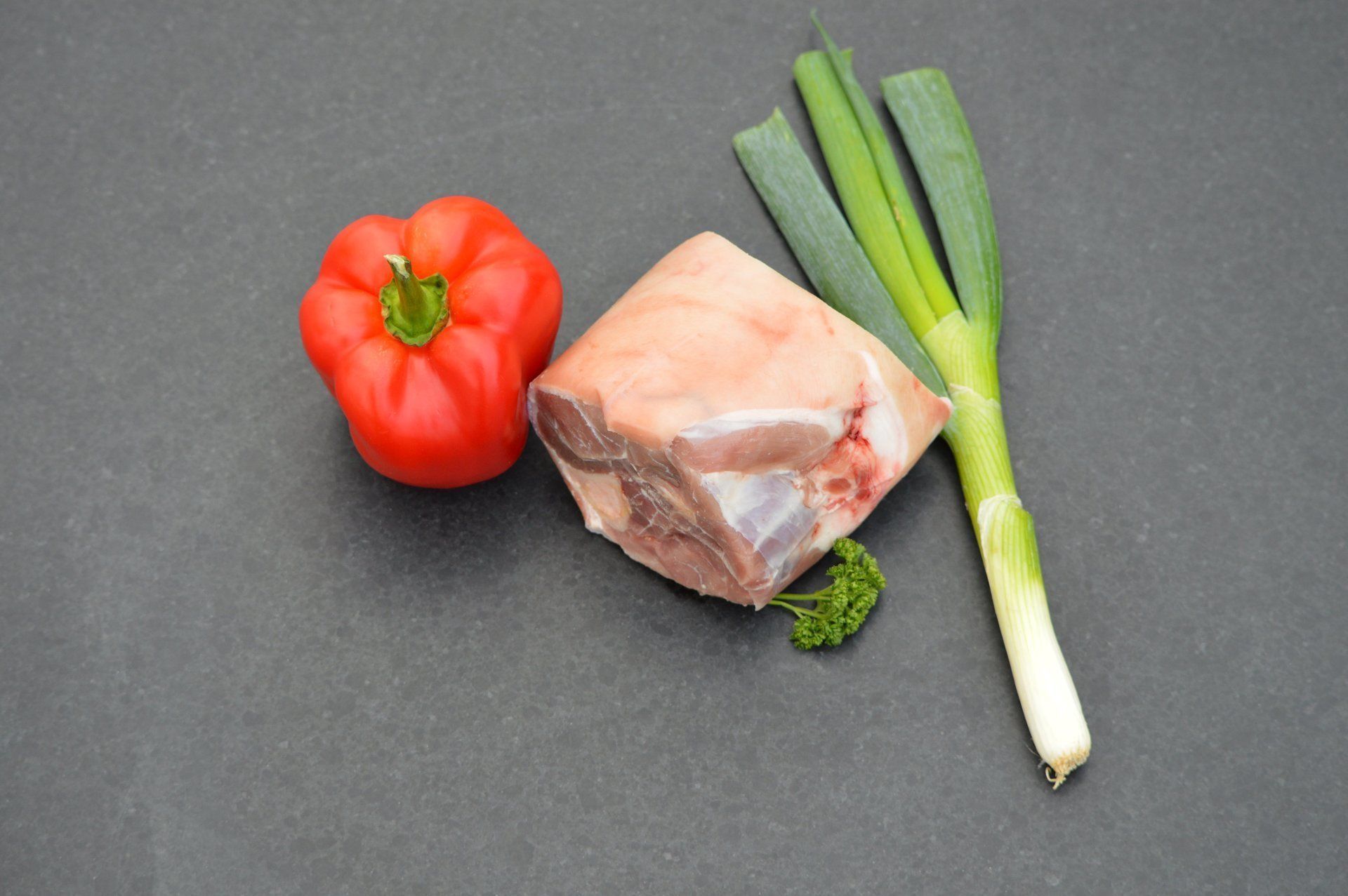 Rauwe varkenschouder, rode paprika en groene ui op een donker oppervlak.