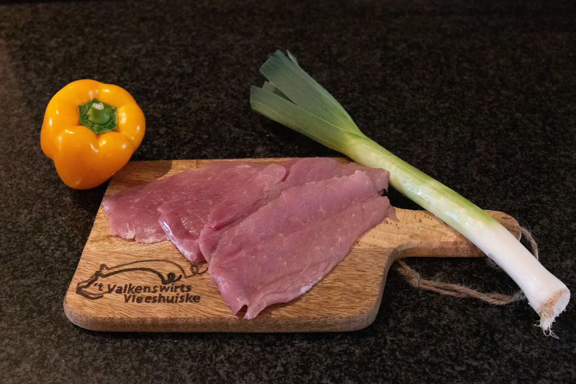 Gele paprika, gesneden vlees op een houten plank, prei op een donker aanrecht.