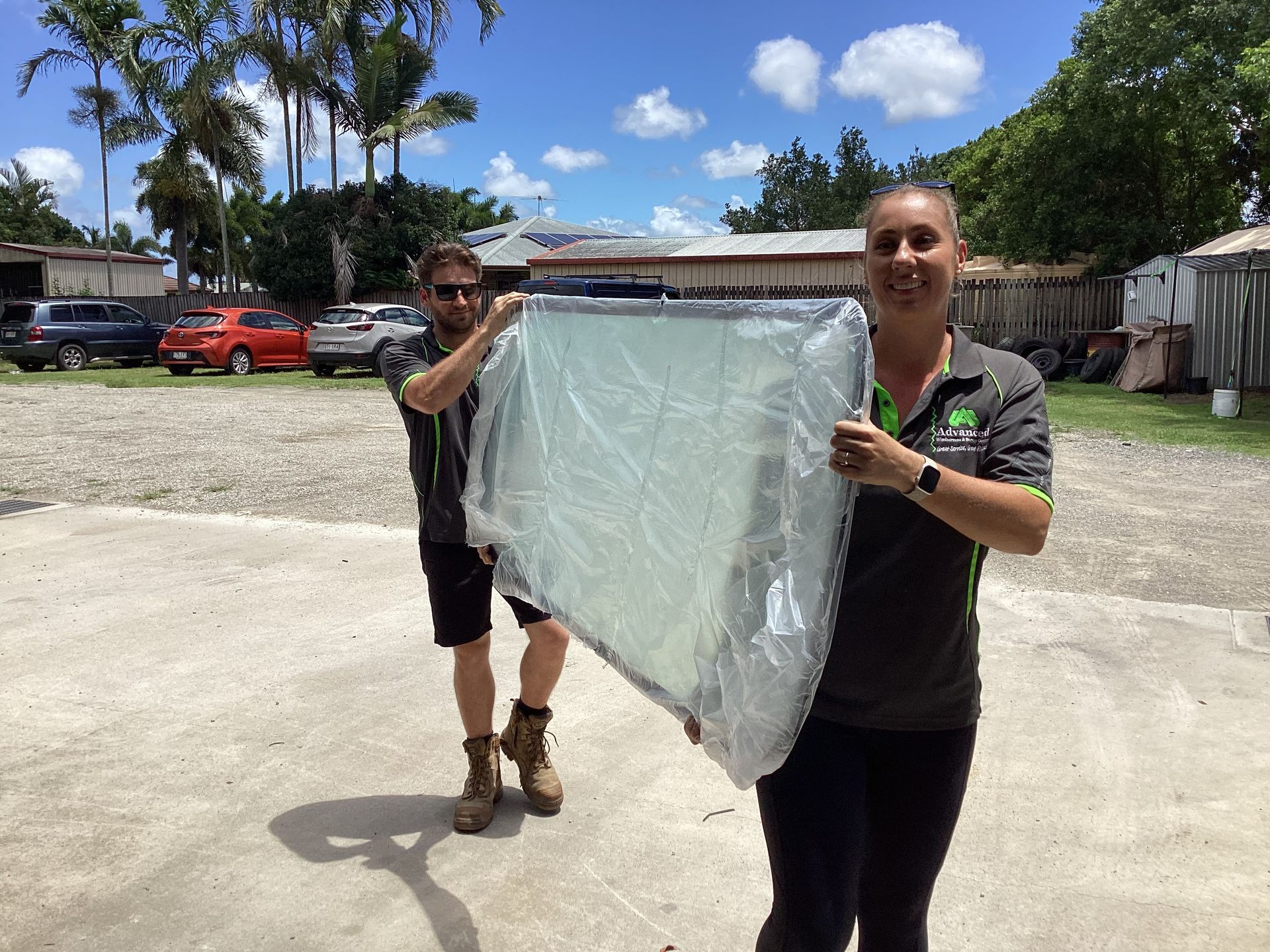 A Man & A Woman Holding Up A New Windscreen In A Plastic Covers— Advanced Windscreens & Service Centre In Proserpine, QLD