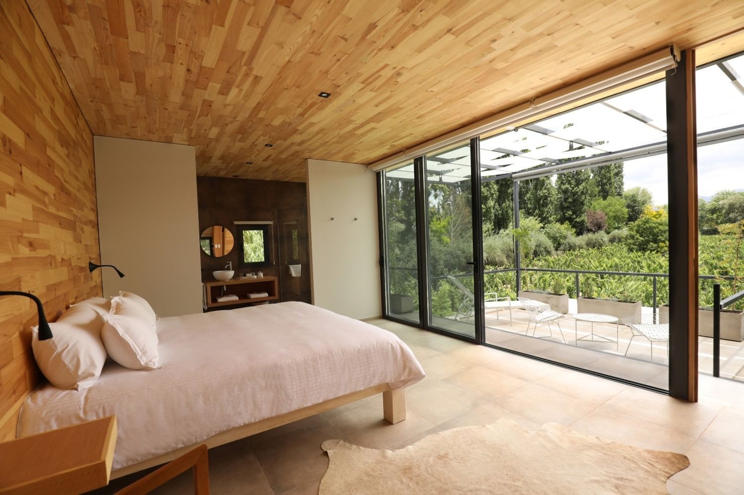 Bedroom with wood walls, ceiling, and floor. Bed with white linens. Large glass doors open to a patio with greenery.