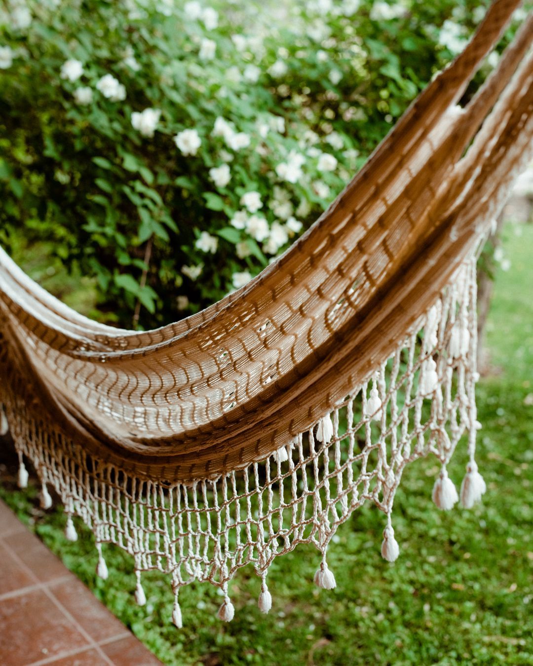 Beige hammock hanging outdoors with white fringe; green foliage in background.