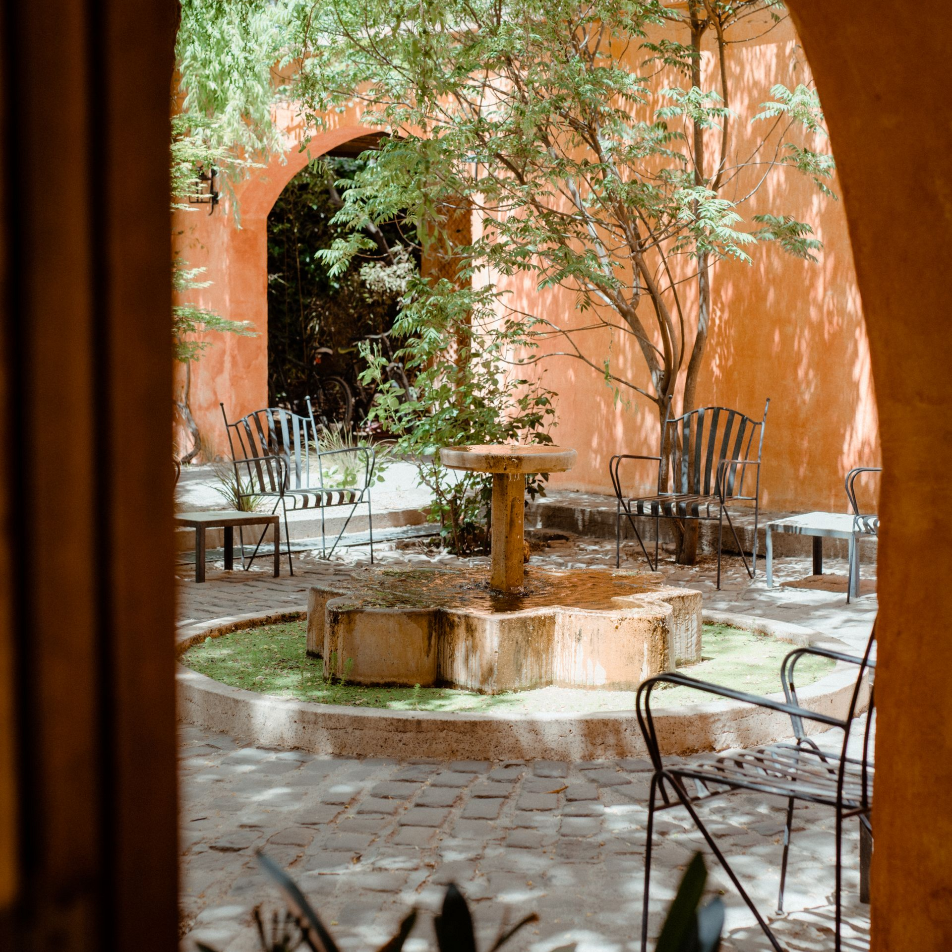 Courtyard with stone fountain, wrought iron chairs, and arched doorway, bathed in sunlight.