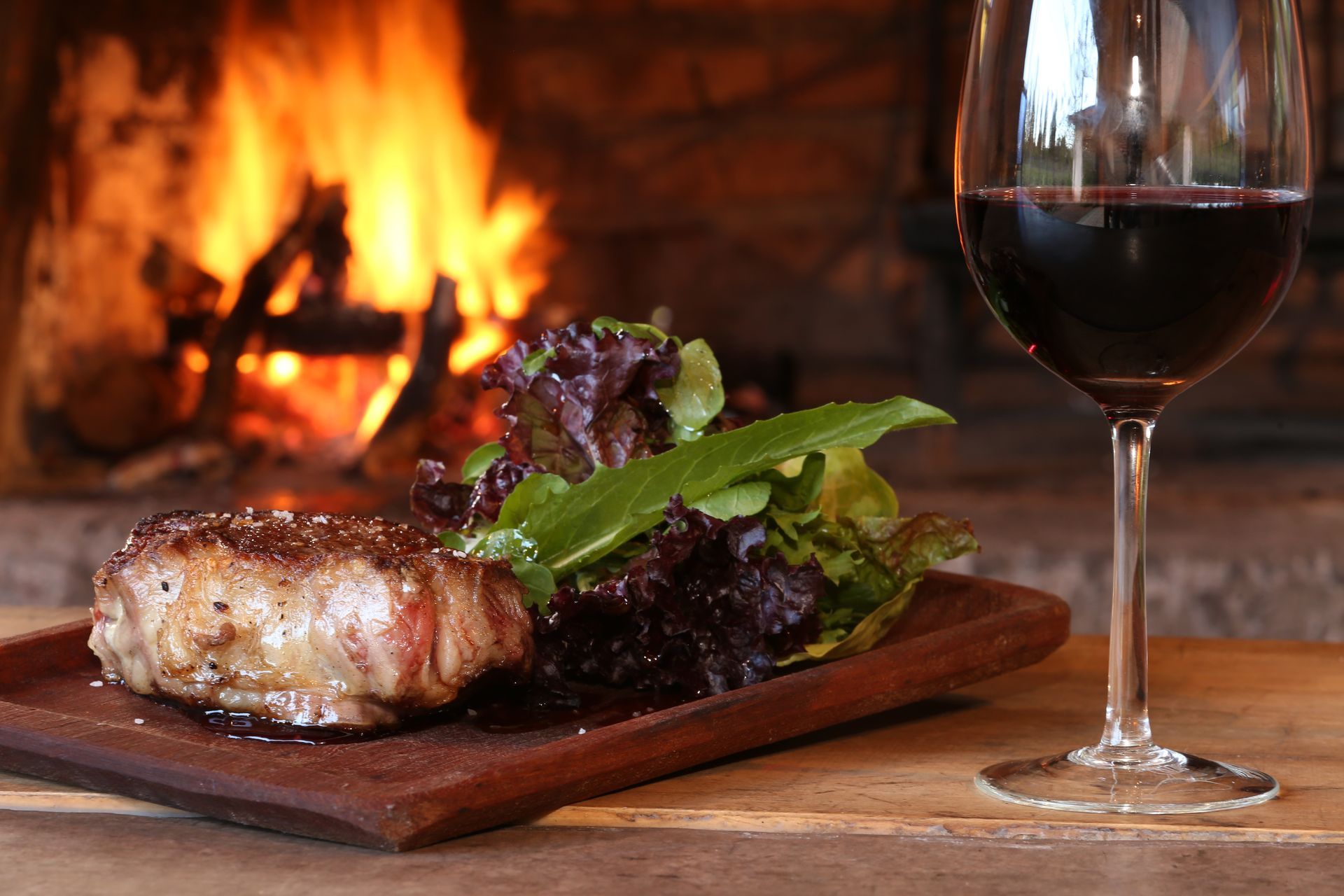 Steak with salad on wooden board, glass of red wine, fireplace in background.