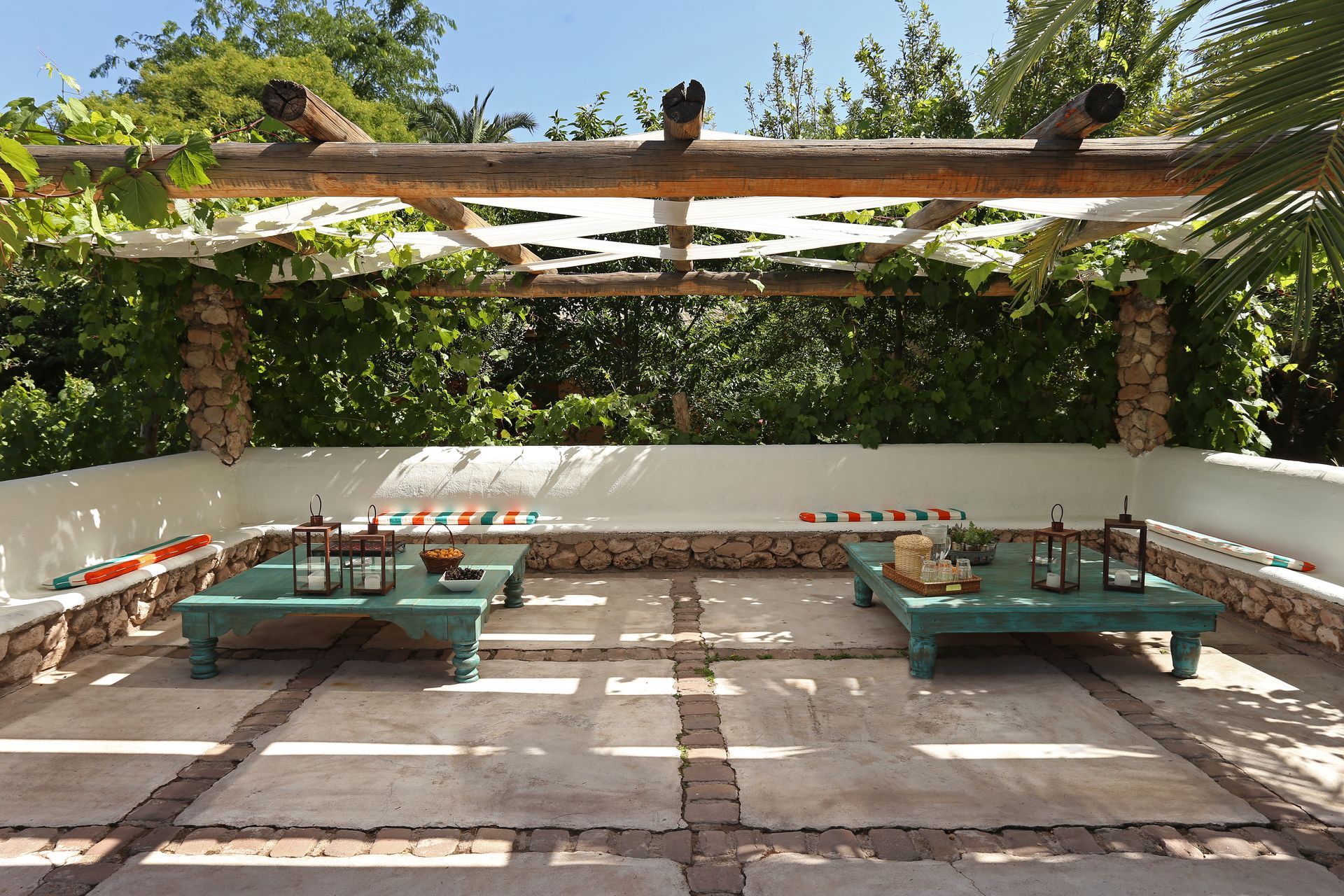Outdoor patio with turquoise tables under a wooden pergola, surrounded by stone walls and lush greenery.