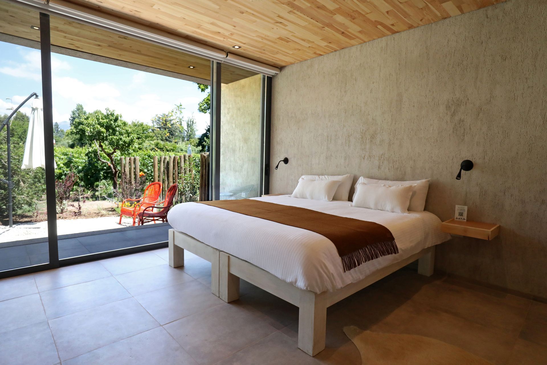 Bedroom with a bed, open glass doors, and a patio with seating; natural textures and light.