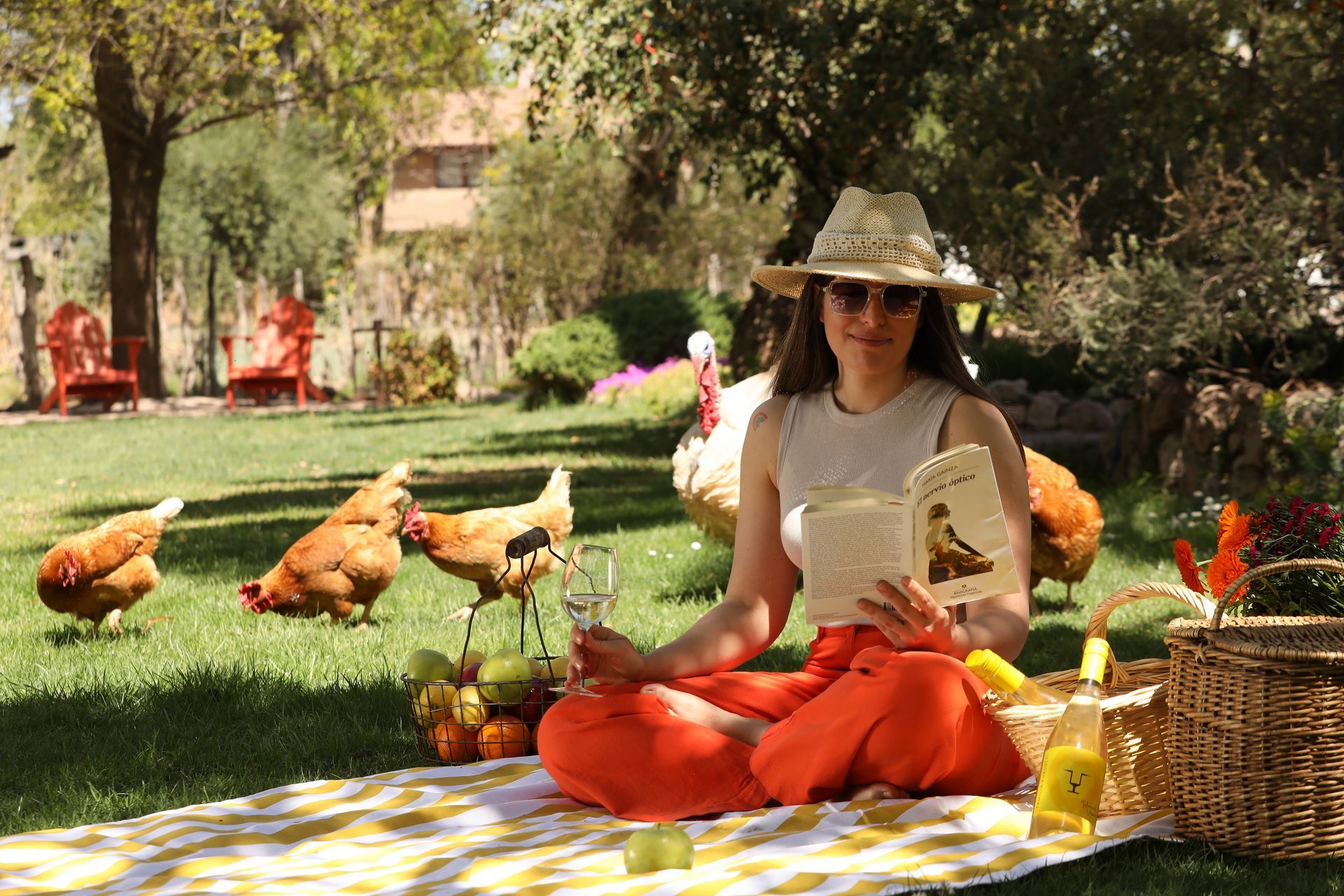 Woman in orange pants reads a book with chickens, fruit, and wine on a picnic blanket outdoors.