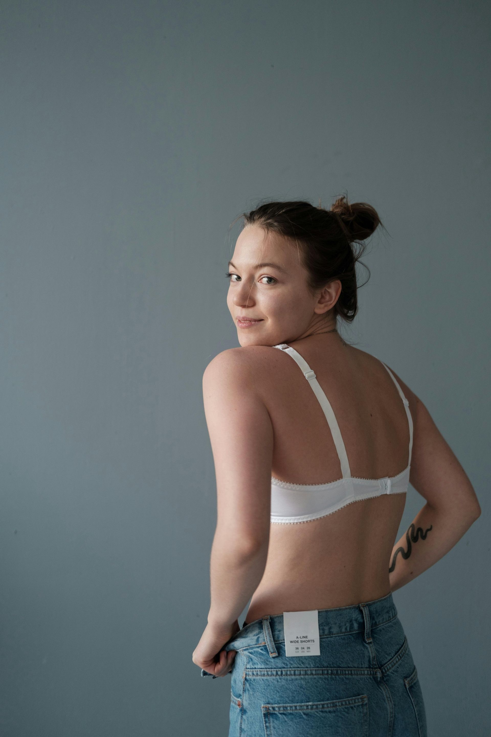 Woman in a white bra and jeans looking over her shoulder against a gray backdrop
