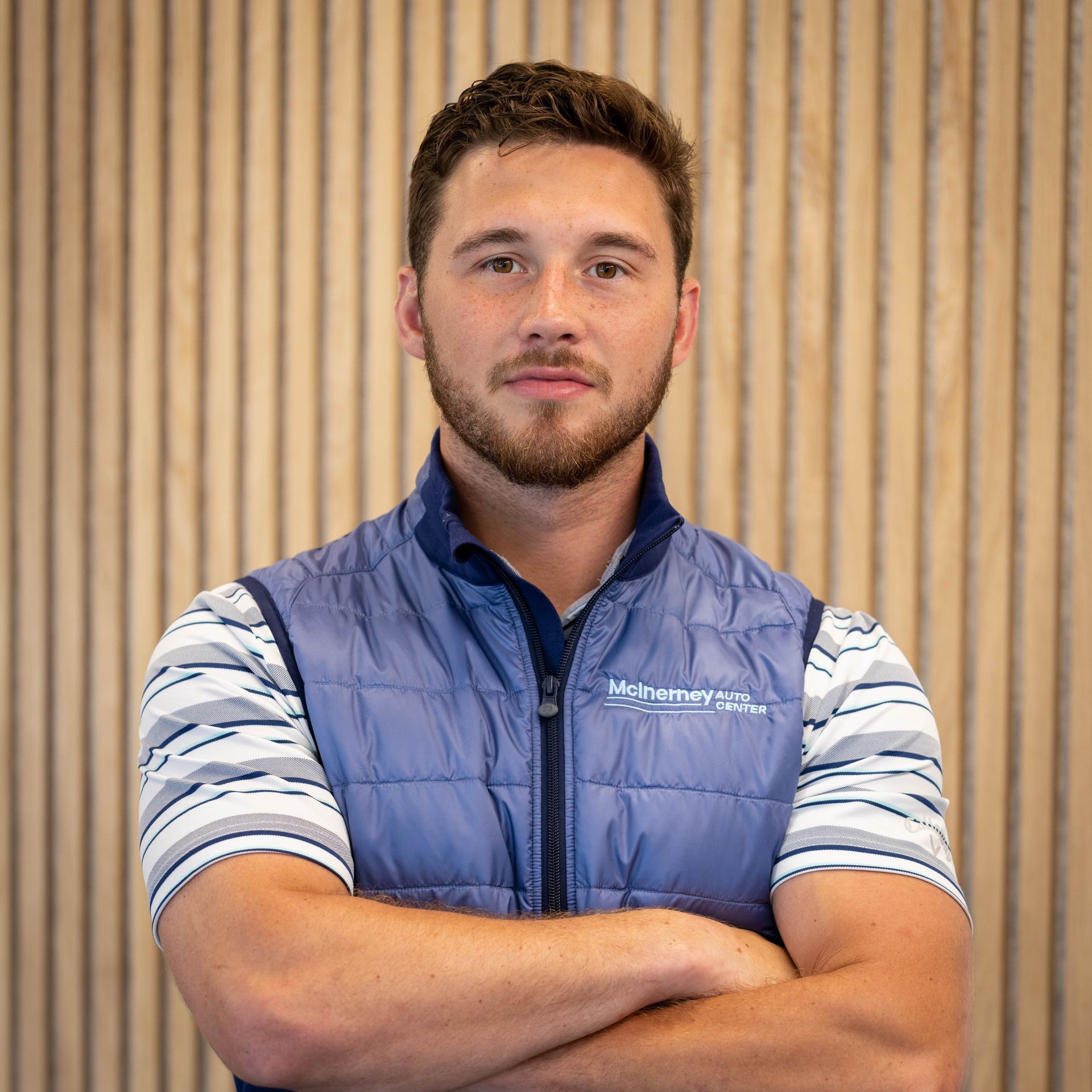 Man with arms crossed, wearing a blue vest over a striped shirt, standing in front of wood paneling. | McInerney Auto Center