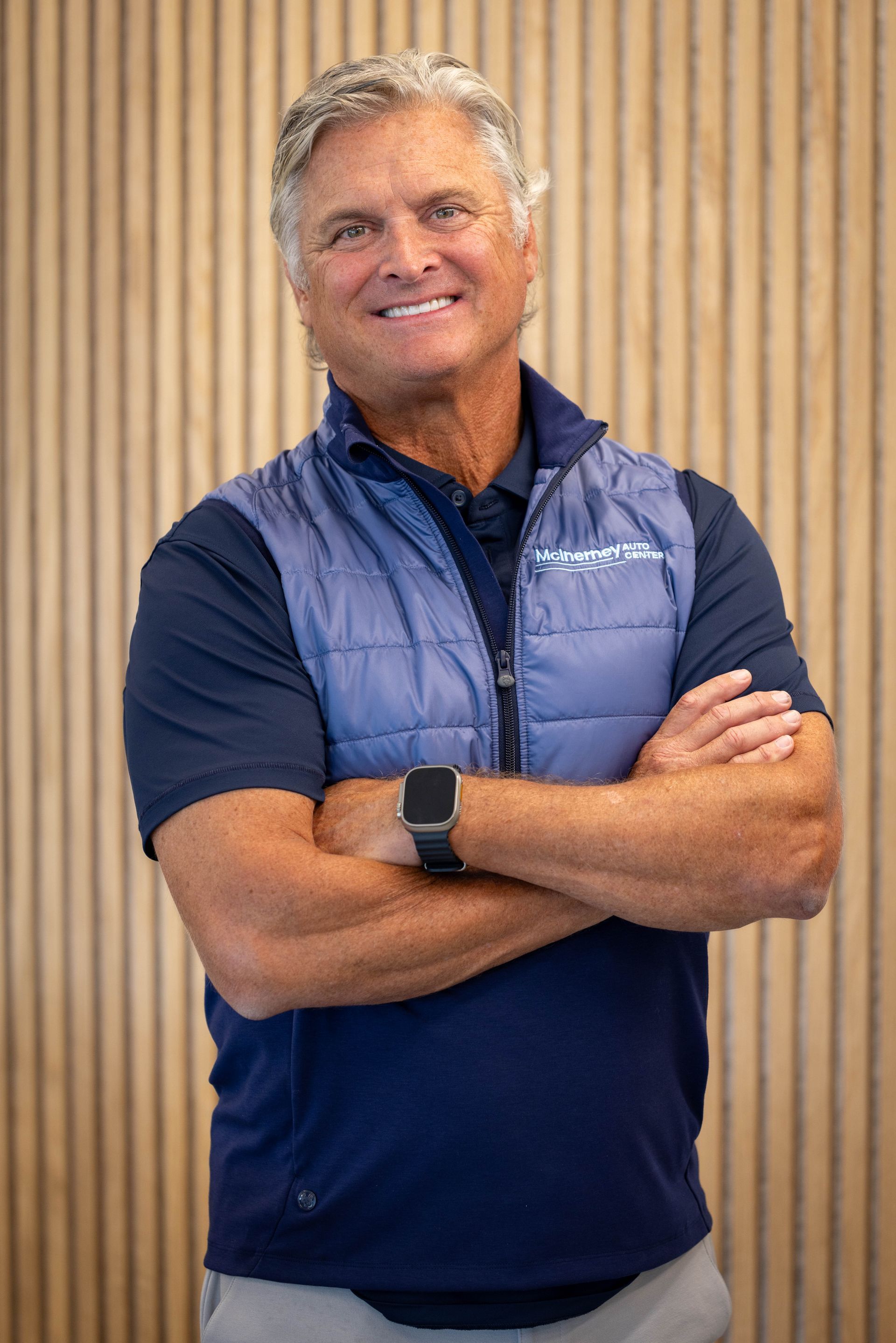 Man with crossed arms wearing a navy vest and polo, smiling. Against a wood-paneled wall. | McInerney Auto Center
