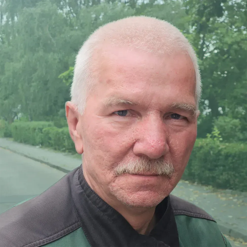 Mann mittleren Alters mit kurzen Haaren und Schnurrbart, der ein grün-braunes Hemd trägt und draußen in die Kamera blickt.