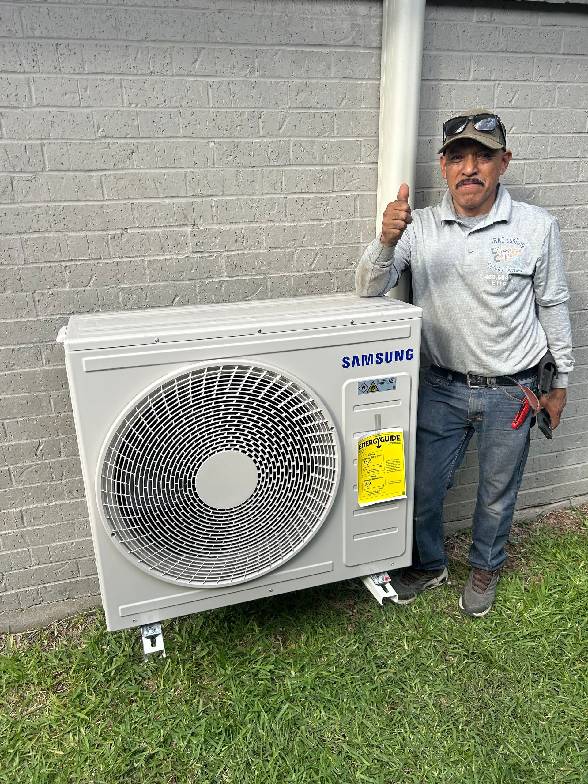 Man gives thumbs up next to a Samsung air conditioning unit. Outdoors, on grass, by a wall.