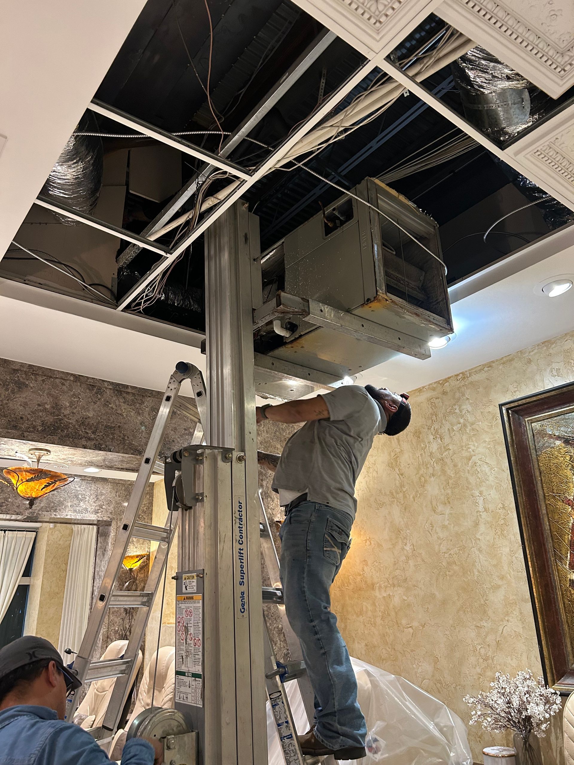 HVAC technician installing an air conditioning unit through a ceiling. He's on a lift, with another worker below.