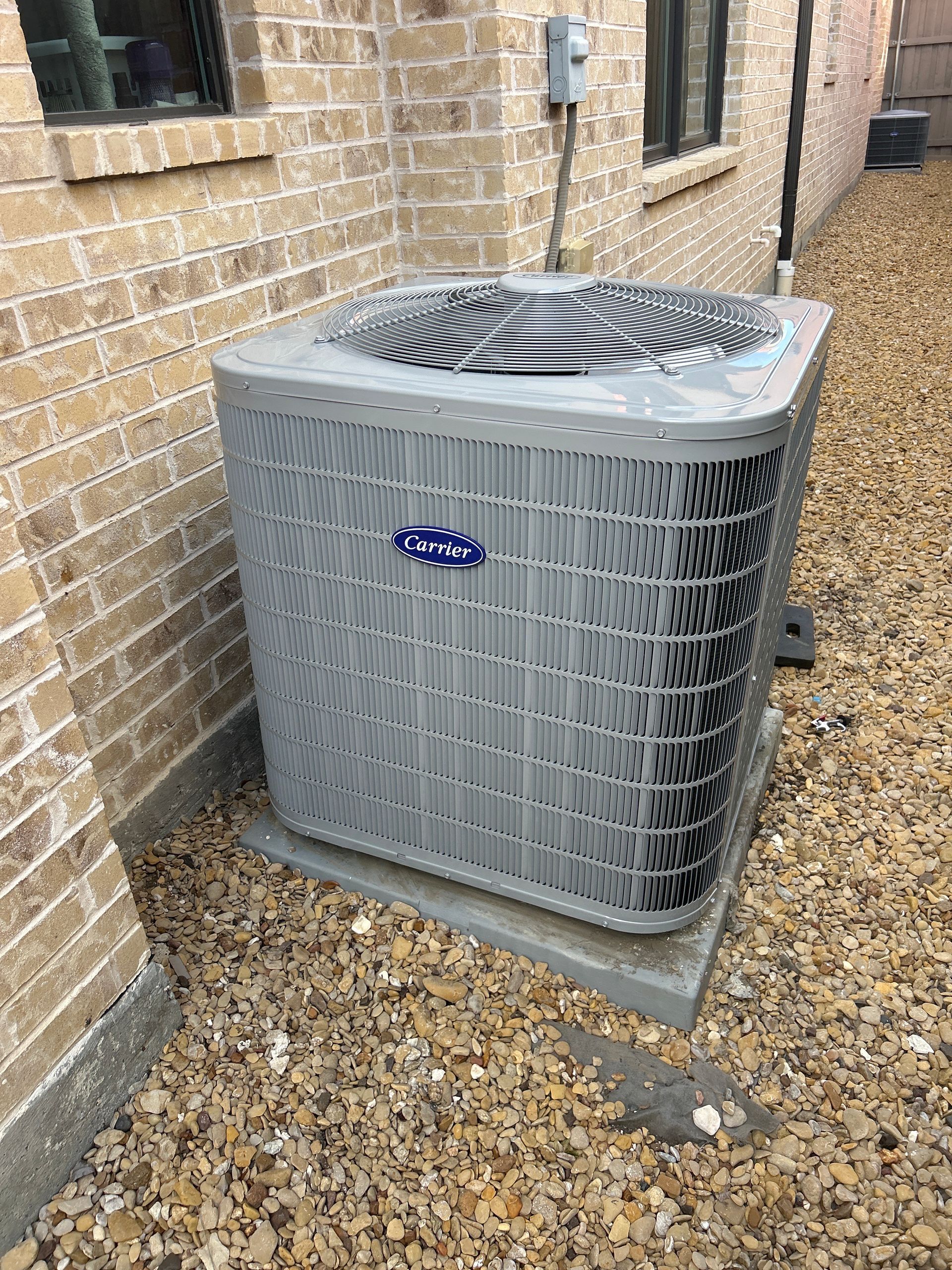 Air conditioner unit, gray, placed next to a brick building on a concrete pad. Gravel surrounds.