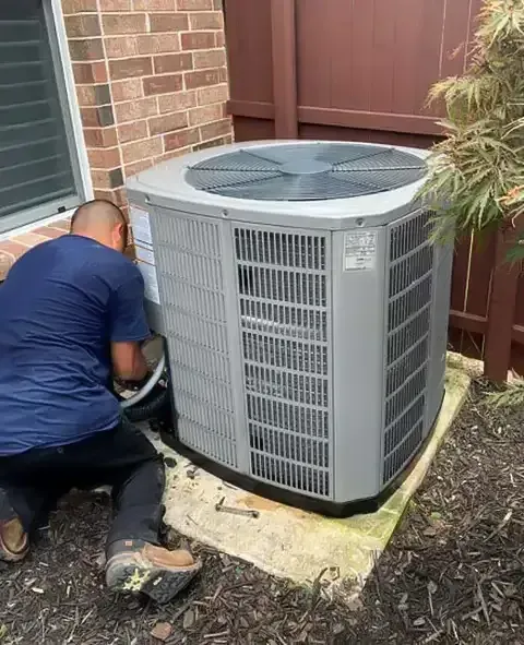 Person working on an air conditioning unit outside near a brick wall and wooden fence.