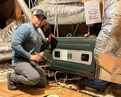 HVAC technician working in an attic, holding a drill, near ductwork and an air handler unit.