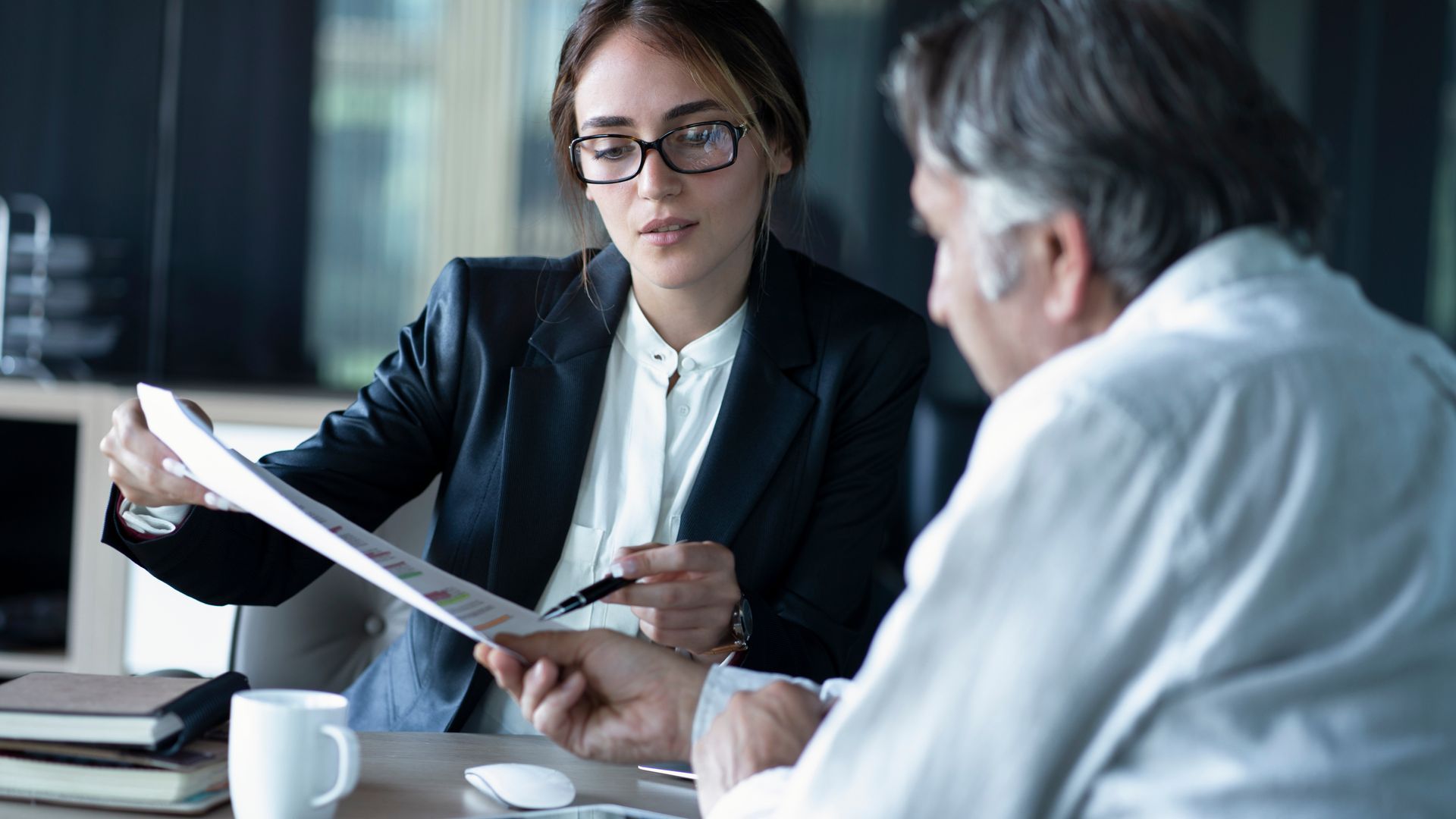 Photograph showing two business professionals in a discussion, with one person handing a document to