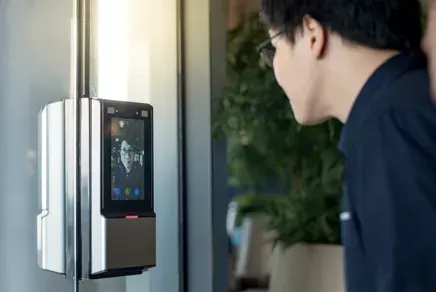 Person using a wall-mounted access control terminal with a touchscreen and camera in a hallway
