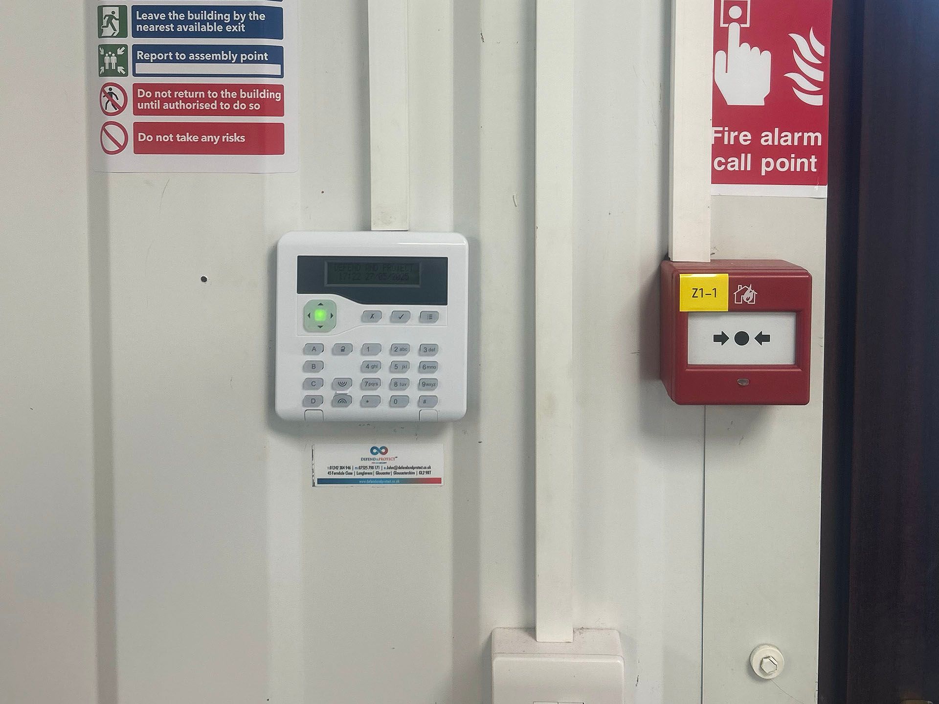 Wall-mounted alarm control panel and red fire alarm call point on a corrugated metal wall.